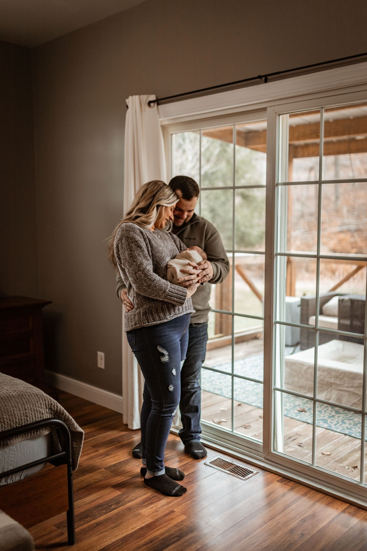 A couple stands together by large glass doors holding their newborn baby in a sunlit room with hardwood floors.