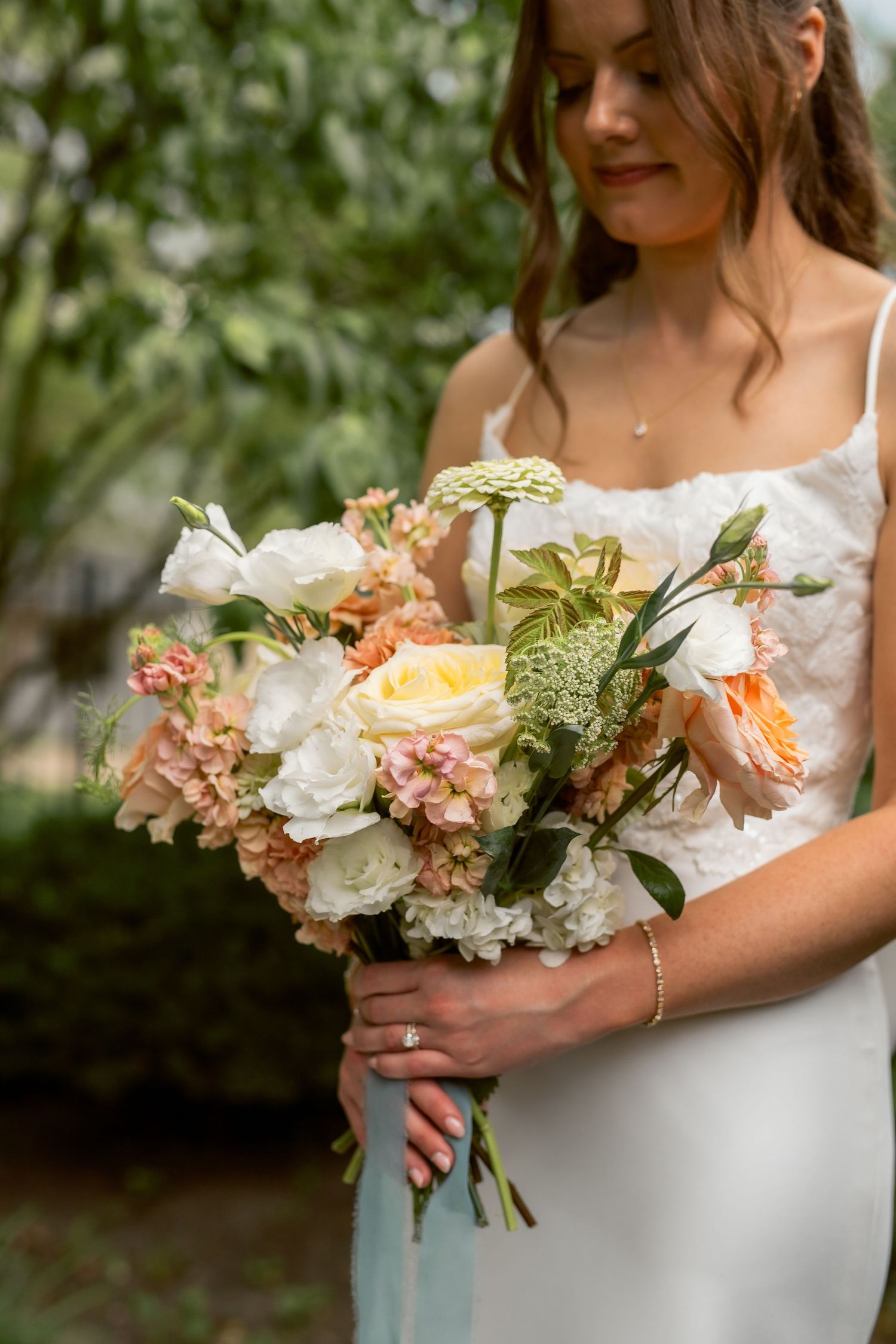 Bride in white gown holding a lush bouquet of peach, cream, and white roses with greenery outdoors.