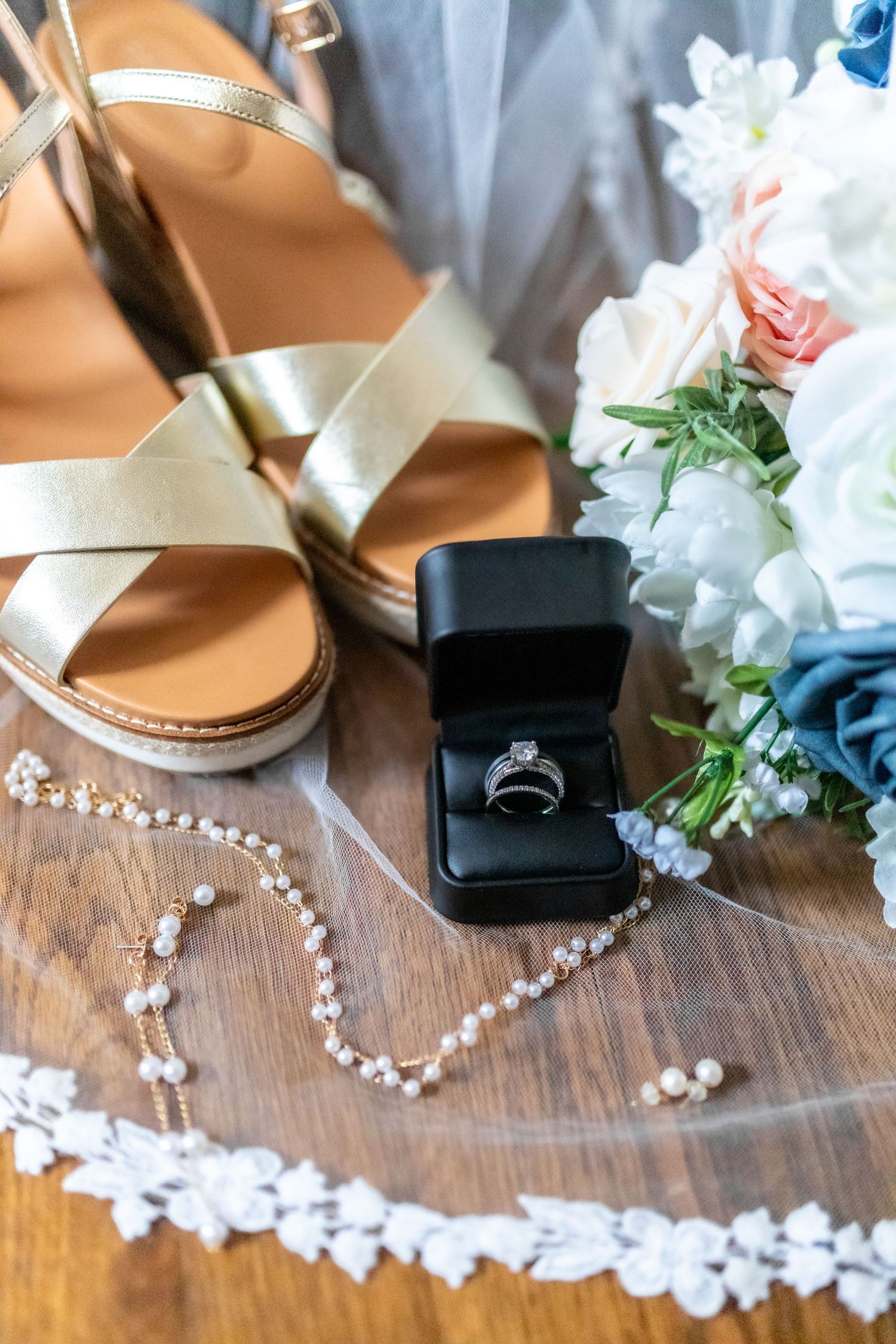 Bridal flat lay featuring gold strappy sandals, diamond ring in black box, pearl necklace, lace veil, and floral bouquet.