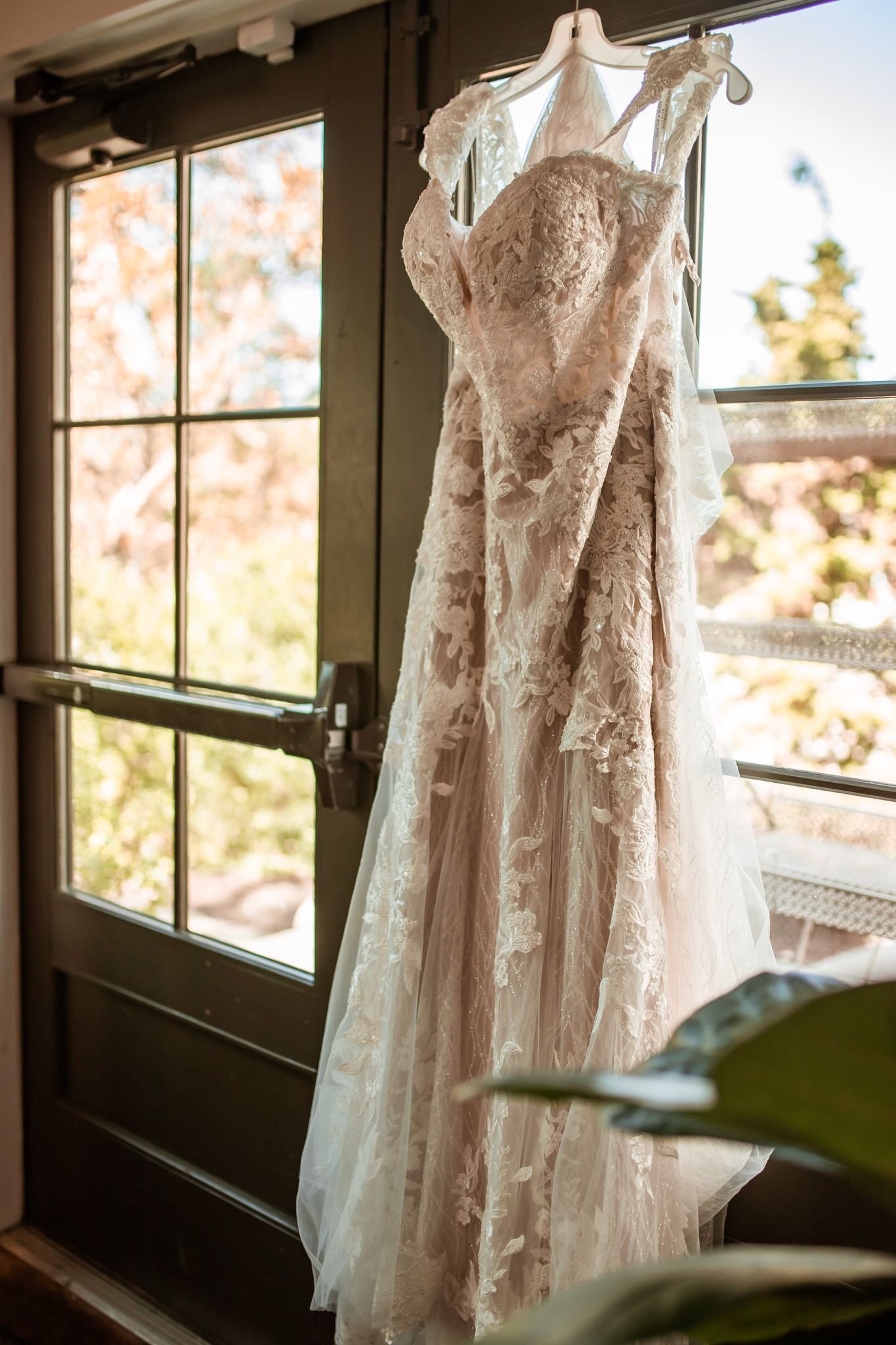 A delicate lace wedding dress with tulle details hangs near a window in natural light, creating a romantic setting.