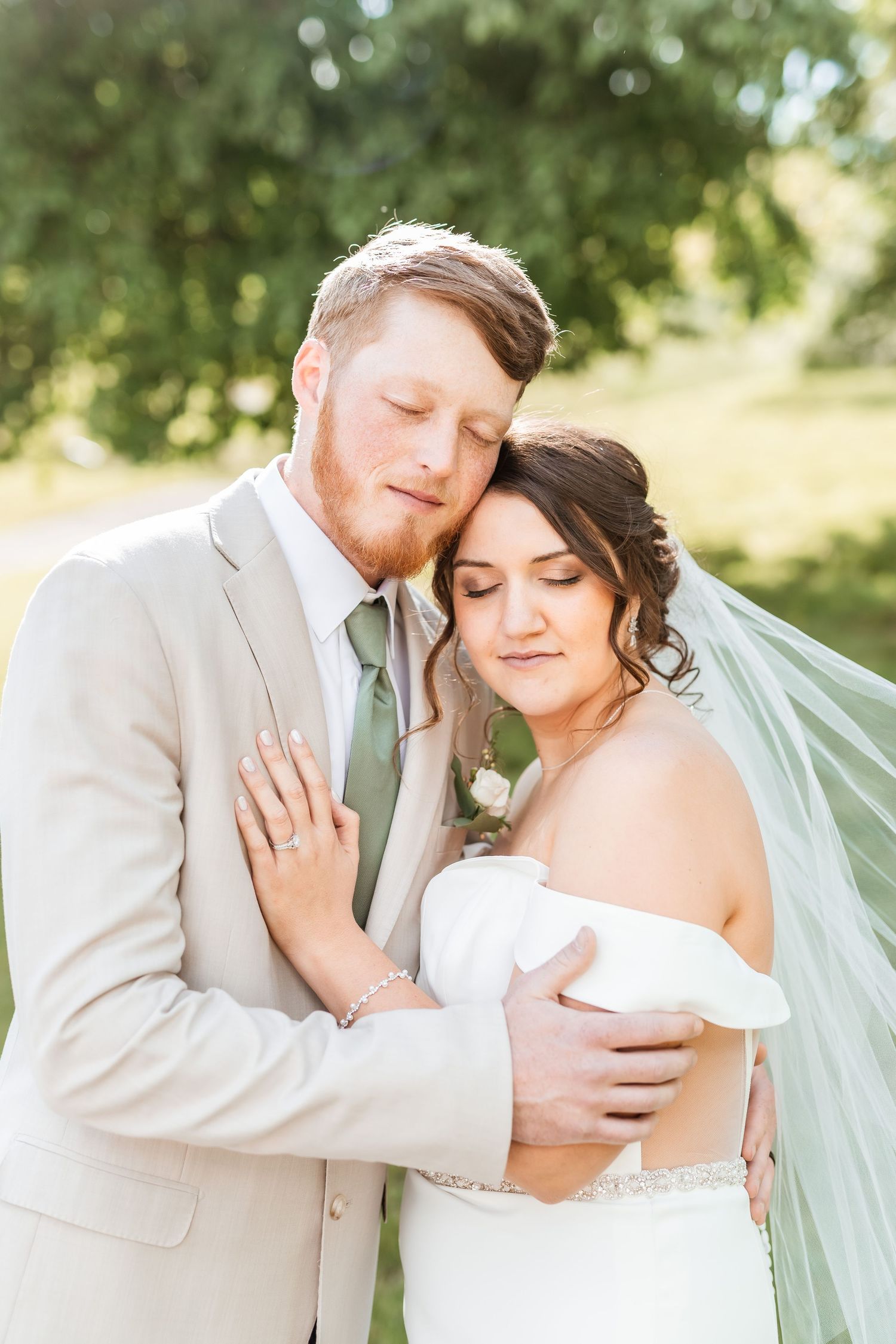 A romantic outdoor wedding photo of newlyweds embracing in soft sunlight, with the bride wearing a veil and off-shoulder gown.
