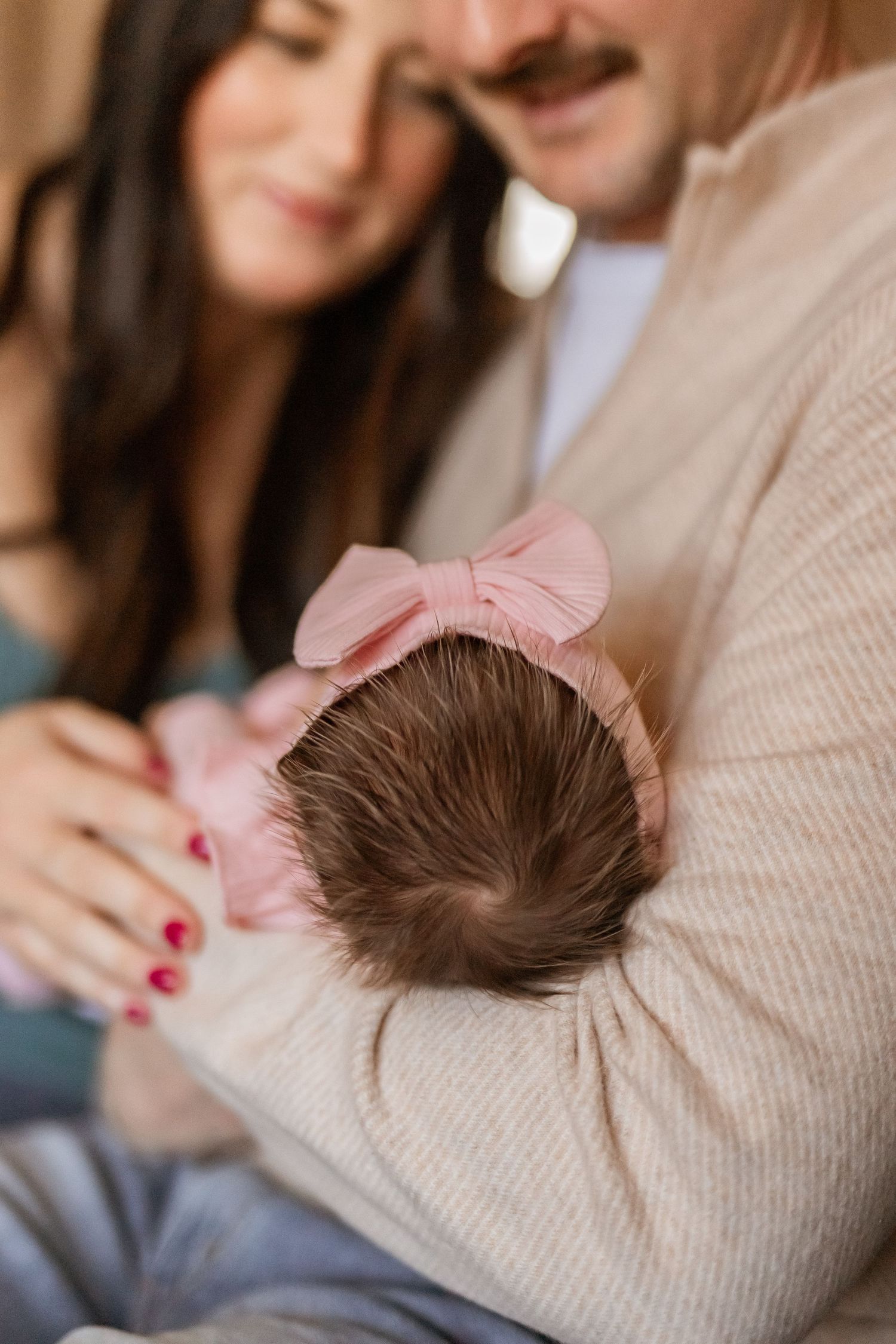 Parents lovingly holding their newborn baby girl wearing a pink bow headband in a warm, tender family portrait.