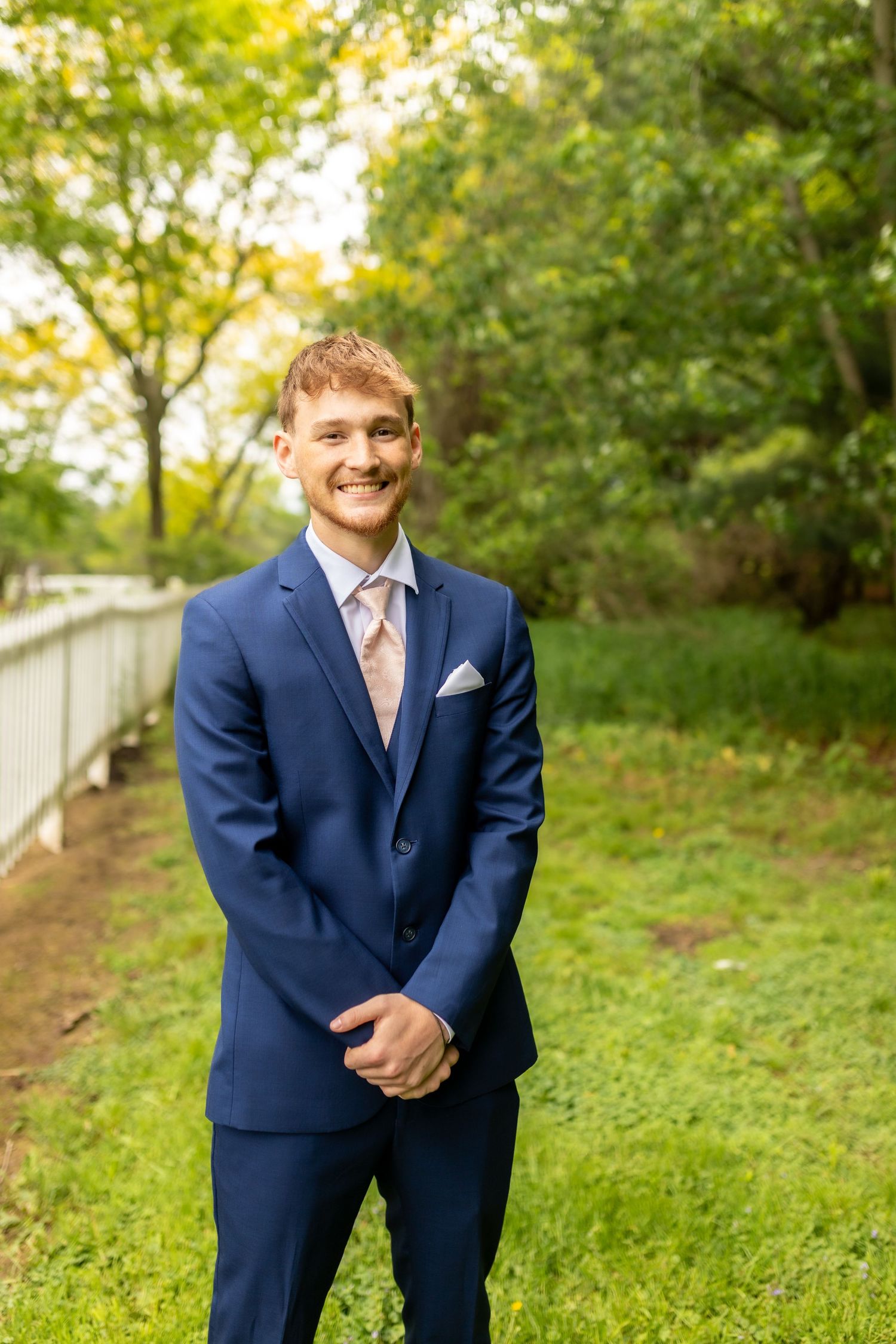 Smiling groom in navy blue suit with pink tie standing outdoors in green garden.