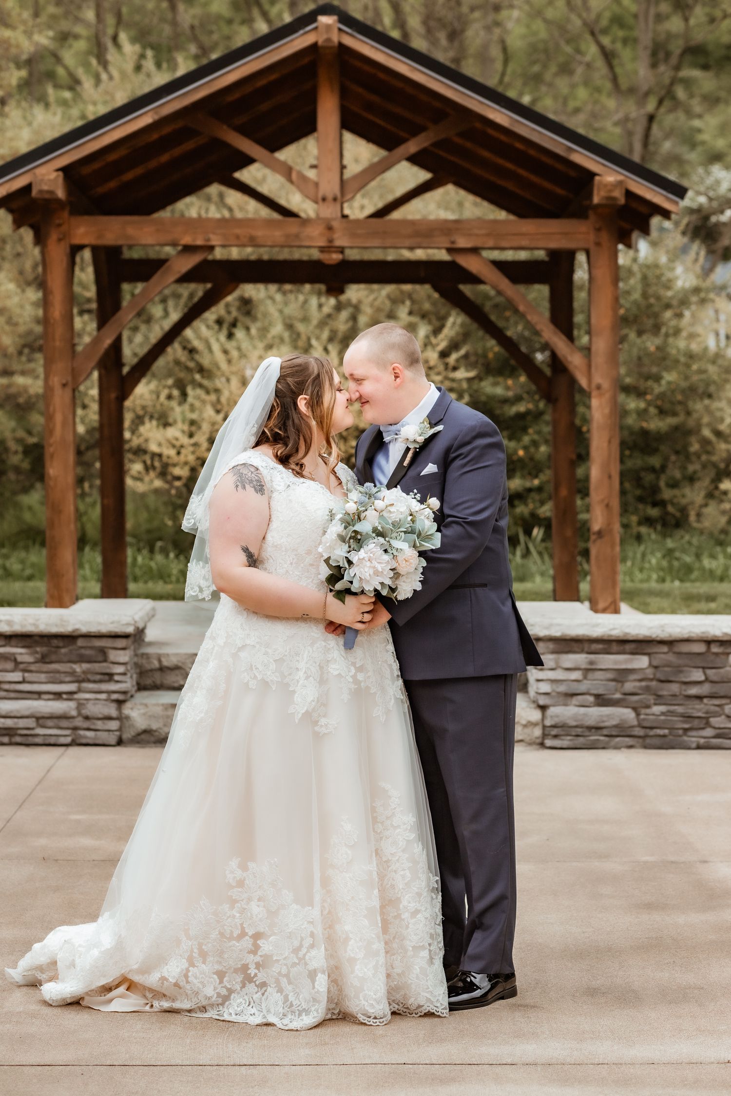 Wedding couple shares a kiss under a rustic wooden pavilion with stone foundation during their ceremony.