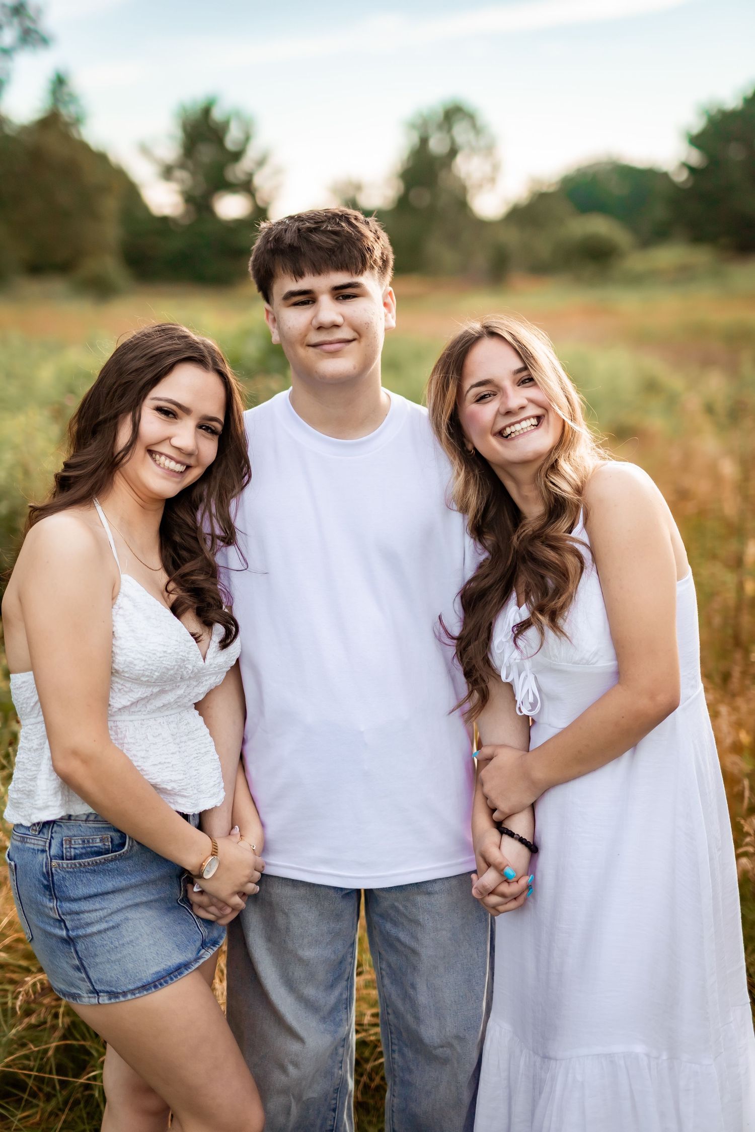 Three people in casual white outfits stand close together smiling during an outdoor photo session in a grassy field at sunset.