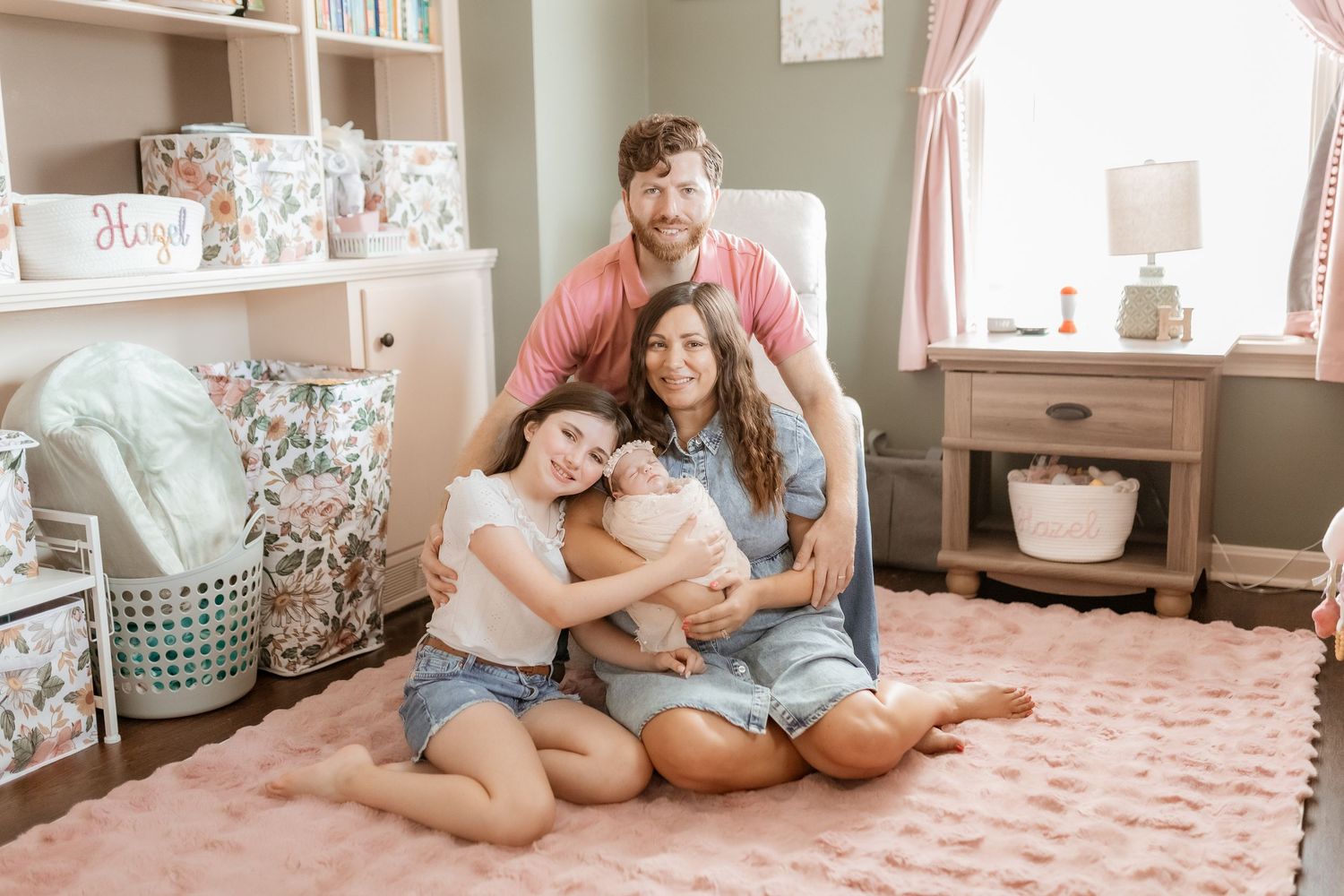 A family sits together on a pink rug in a cozy bedroom with soft decor and natural lighting.