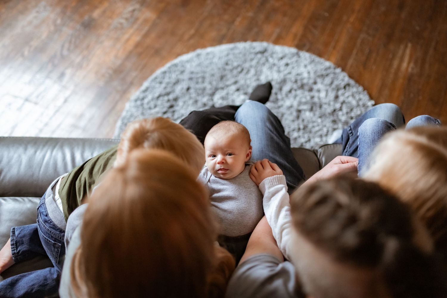 Bird's eye view of family gathering around baby lying on grey rug with hardwood floor visible.
