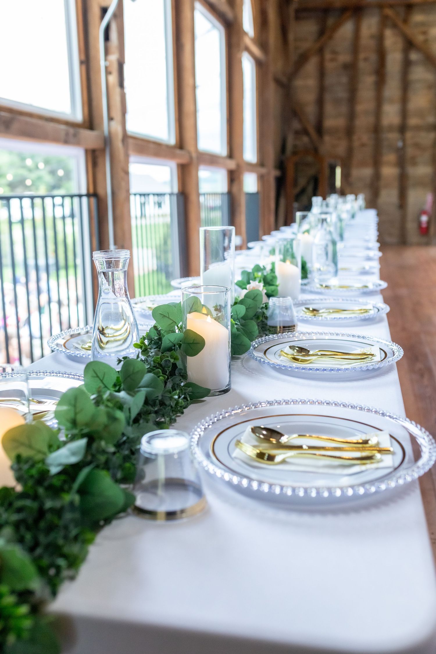 Elegant barn wedding reception table with gold cutlery, white candles, greenery runner, and glass charger plates.