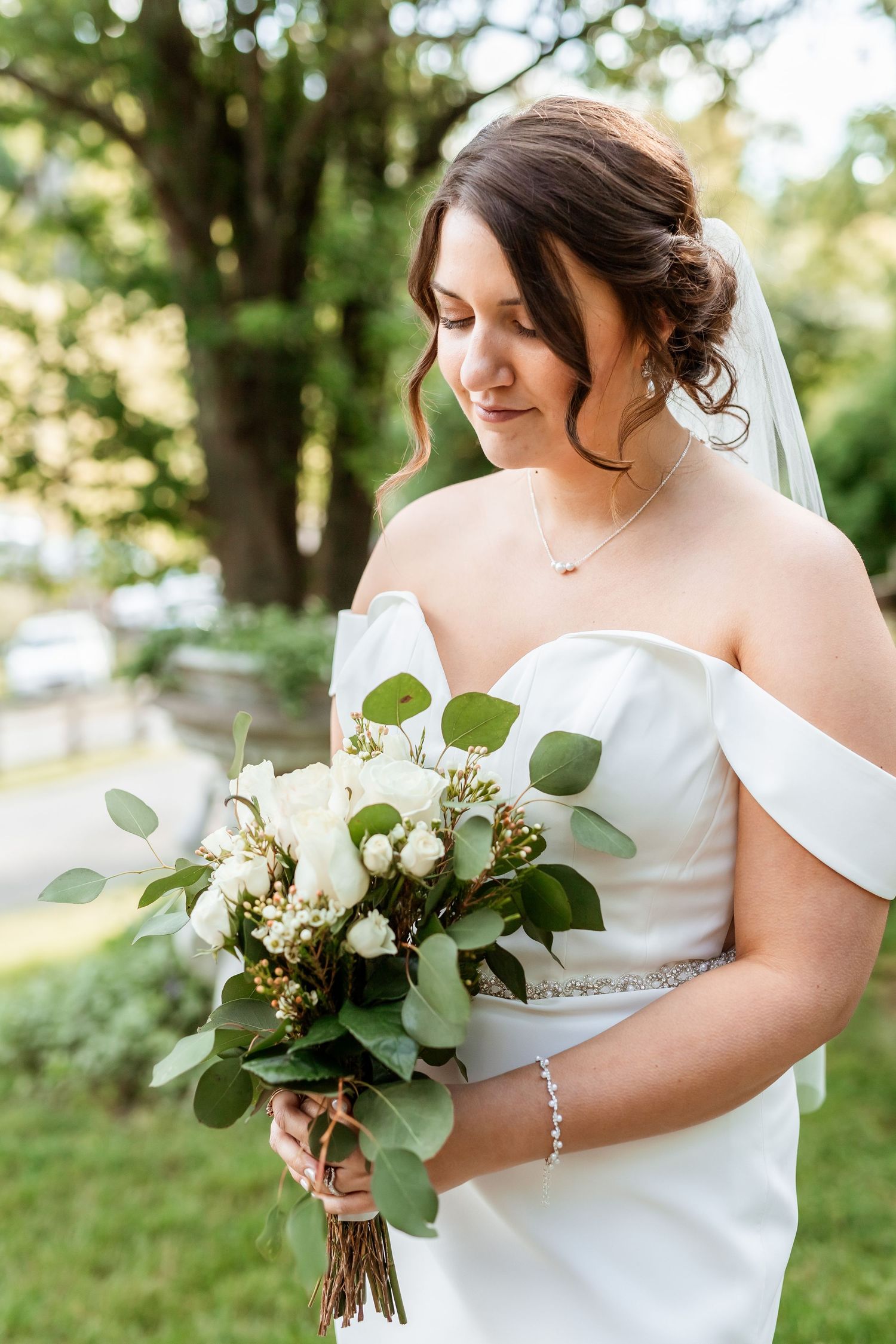 Elegant bride in off-shoulder white gown holds romantic bouquet of white flowers and eucalyptus leaves in outdoor garden setting.