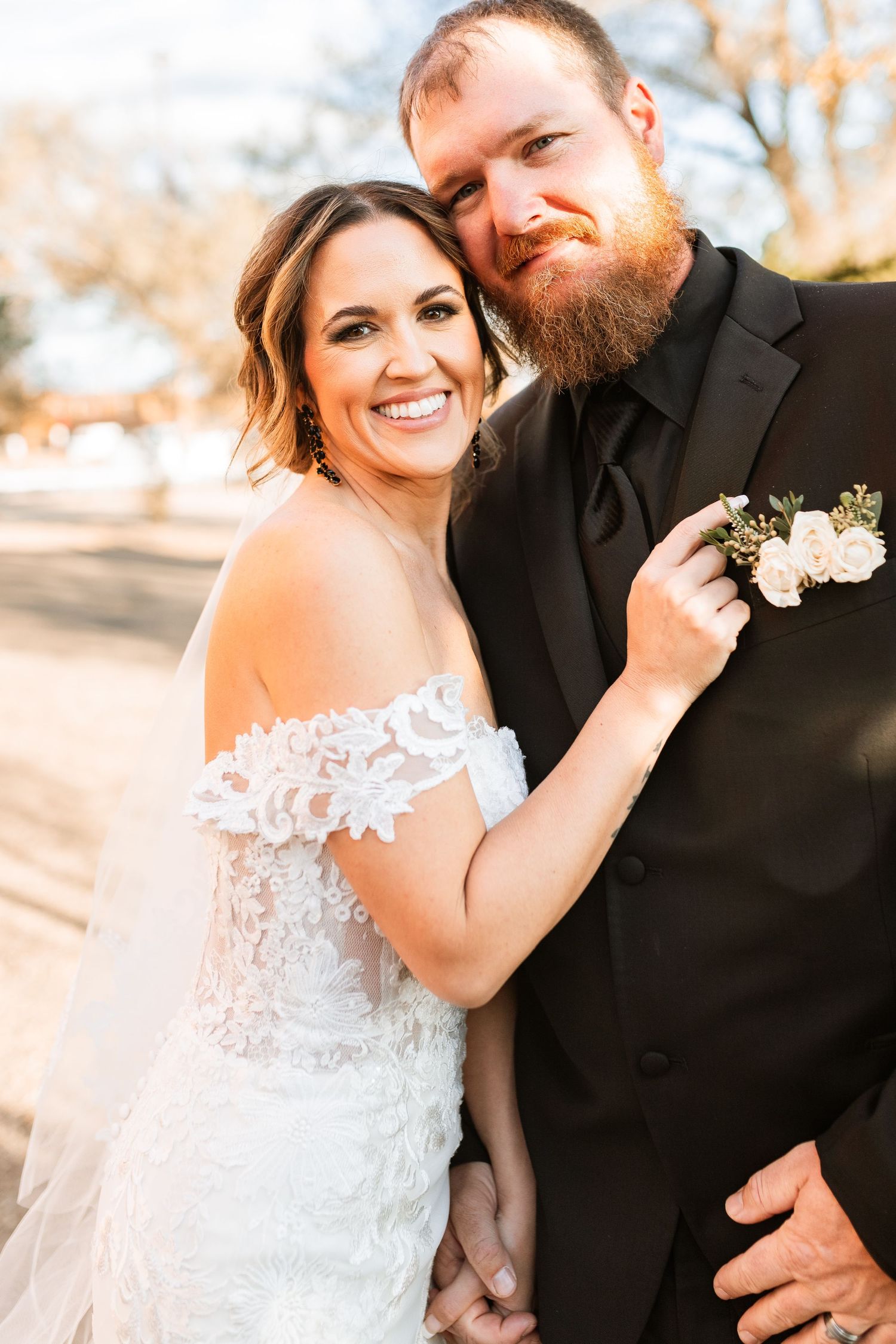 Happy couple poses together outdoors on their wedding day, with the bride in lace and groom in a black suit.