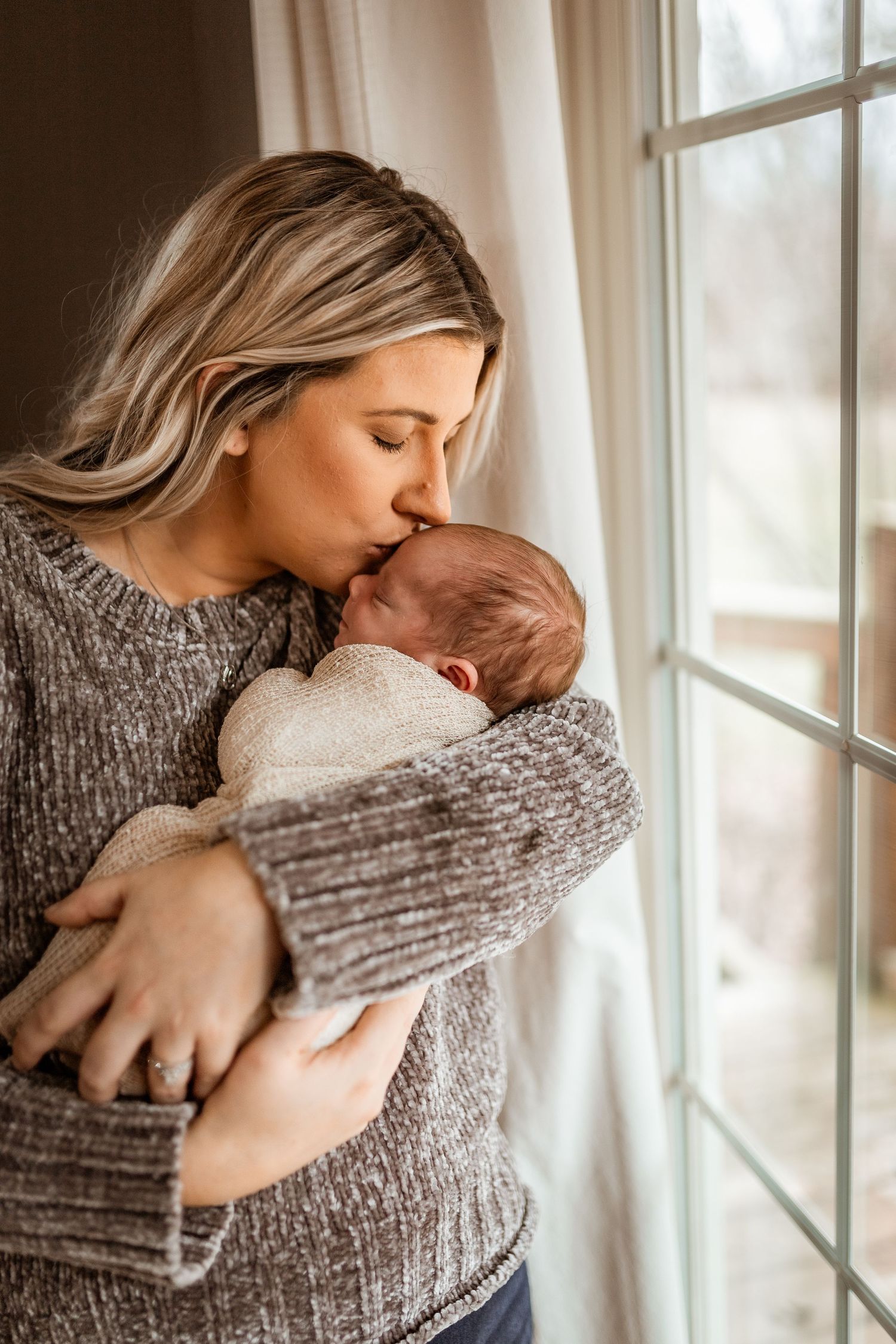 A tender moment by a window as parent in gray sweater cradles and kisses newborn baby wrapped in white blanket.