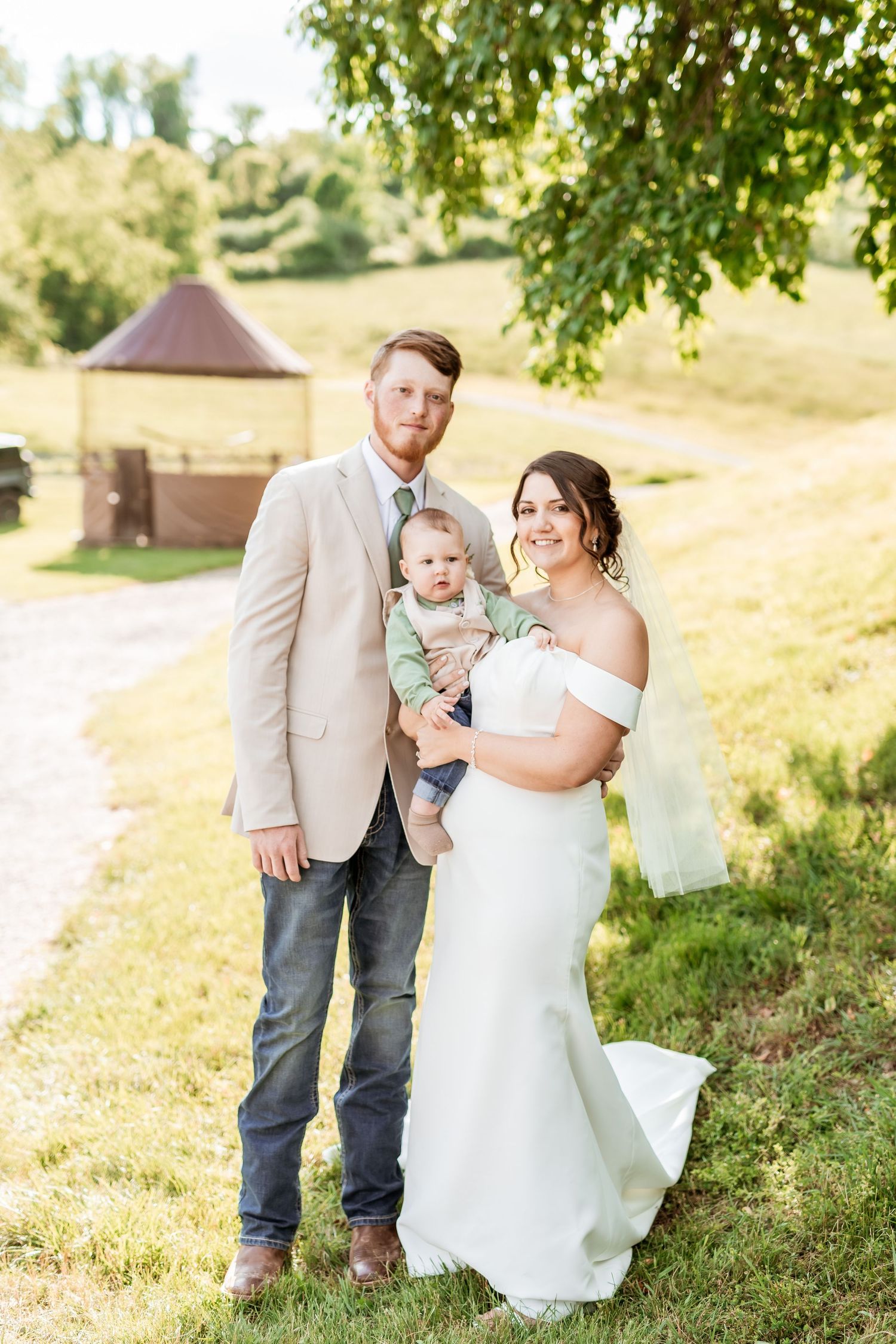A couple and their baby pose for a wedding photo on a sunny hillside with a gazebo in the background.