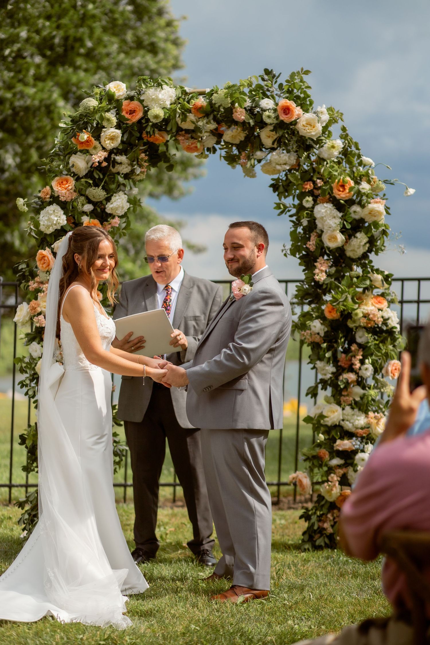 Bride and groom exchanging vows under a floral arch with peach and white flowers at an outdoor wedding ceremony.