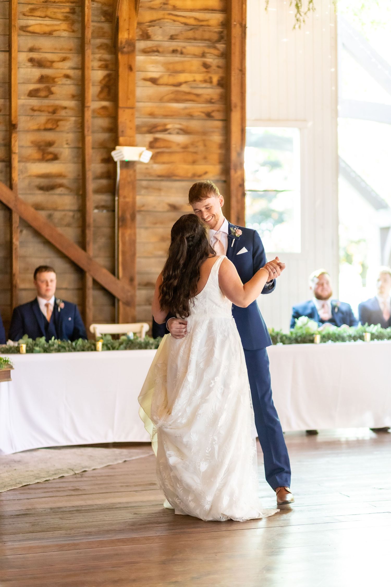 Bride and groom sharing first dance in rustic barn wedding venue with wood walls and greenery decorations.