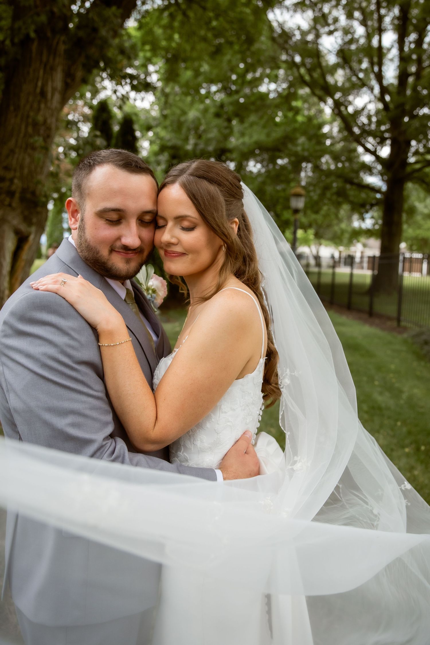 Romantic wedding couple embracing outdoors, bride in white gown with flowing veil and groom in gray suit among green trees.