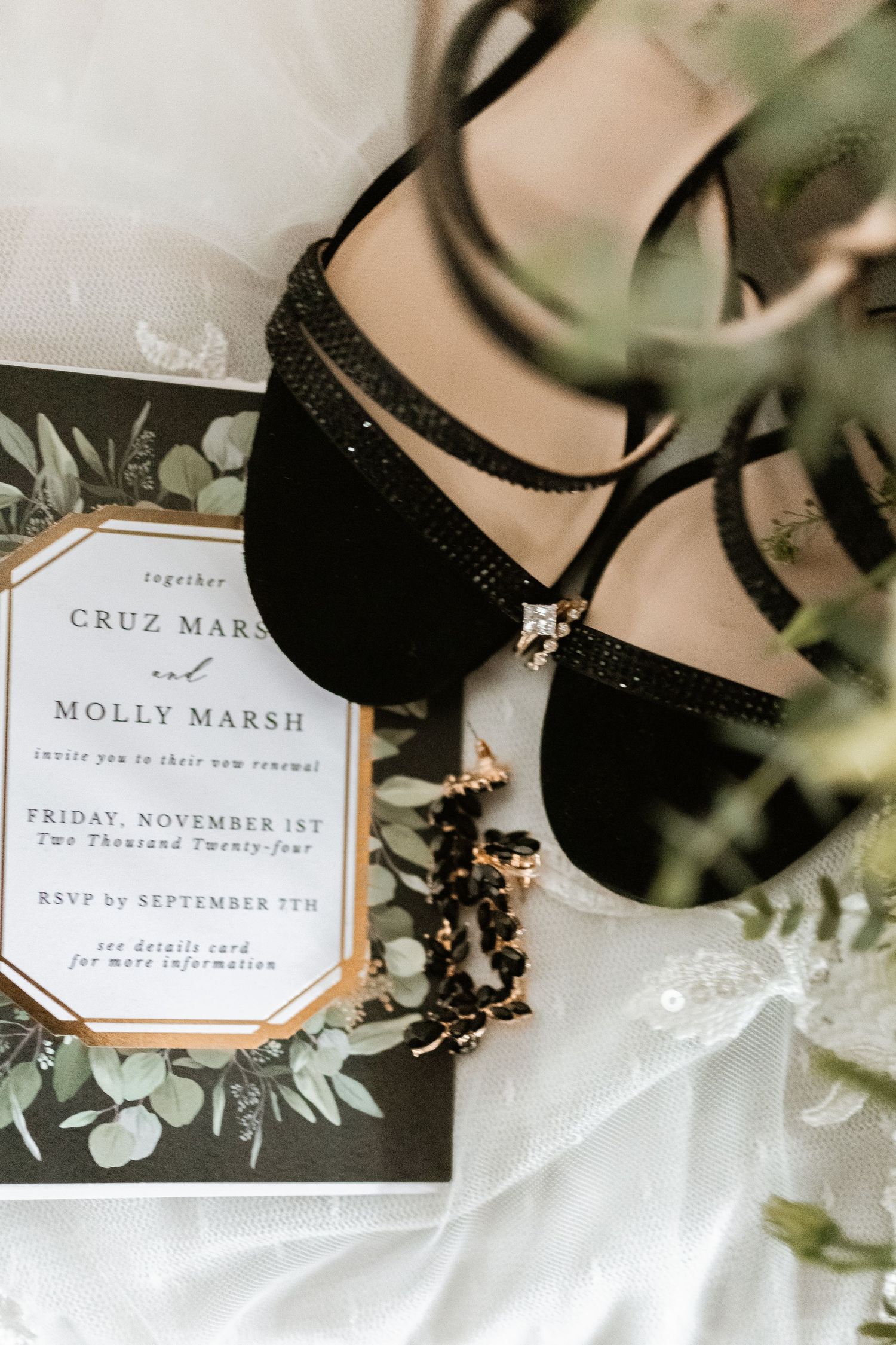 A wedding invitation with black dress shoes photographed alongside greenery and floral details.