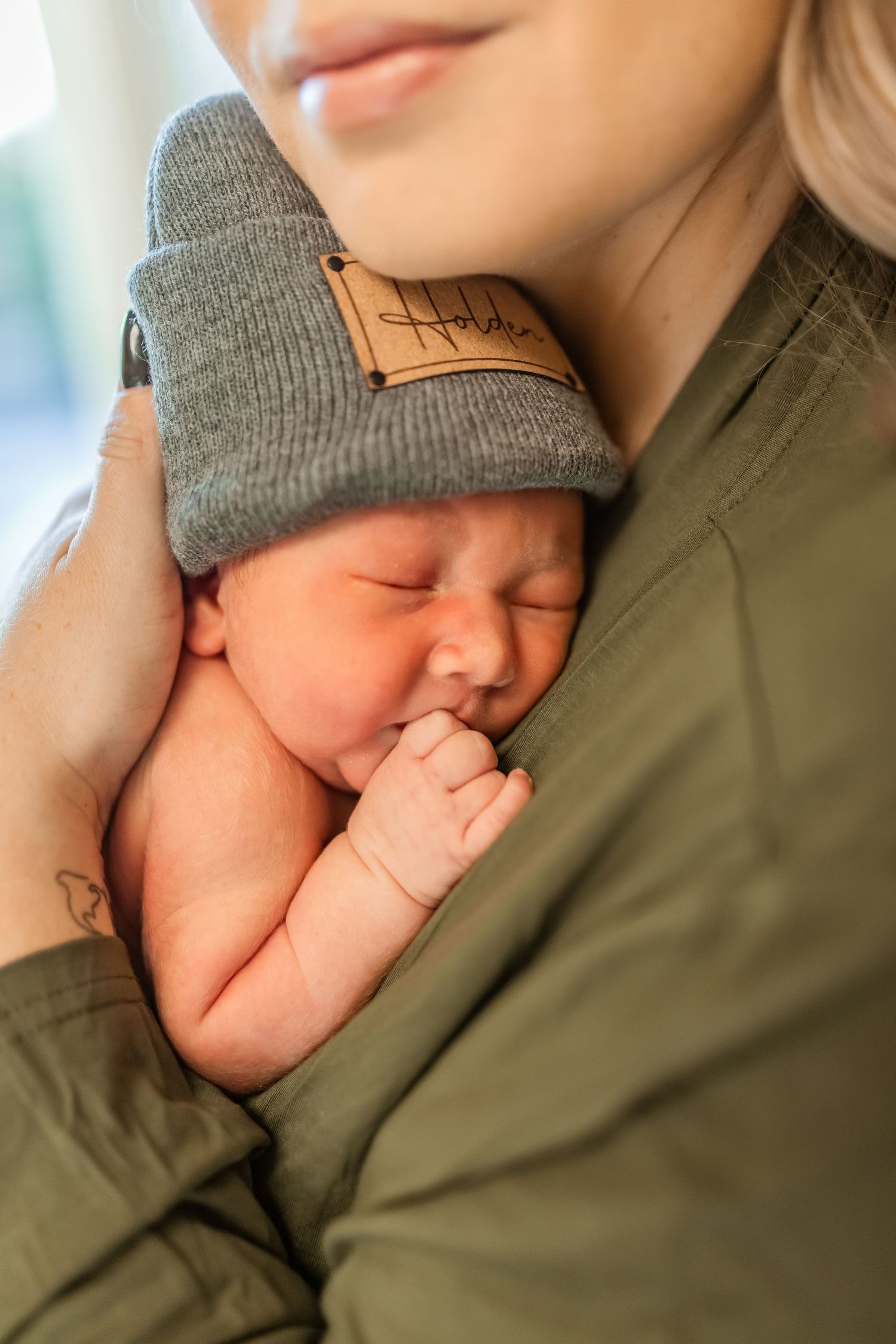 Newborn baby wearing gray beanie sleeps peacefully against parent in olive green clothing.