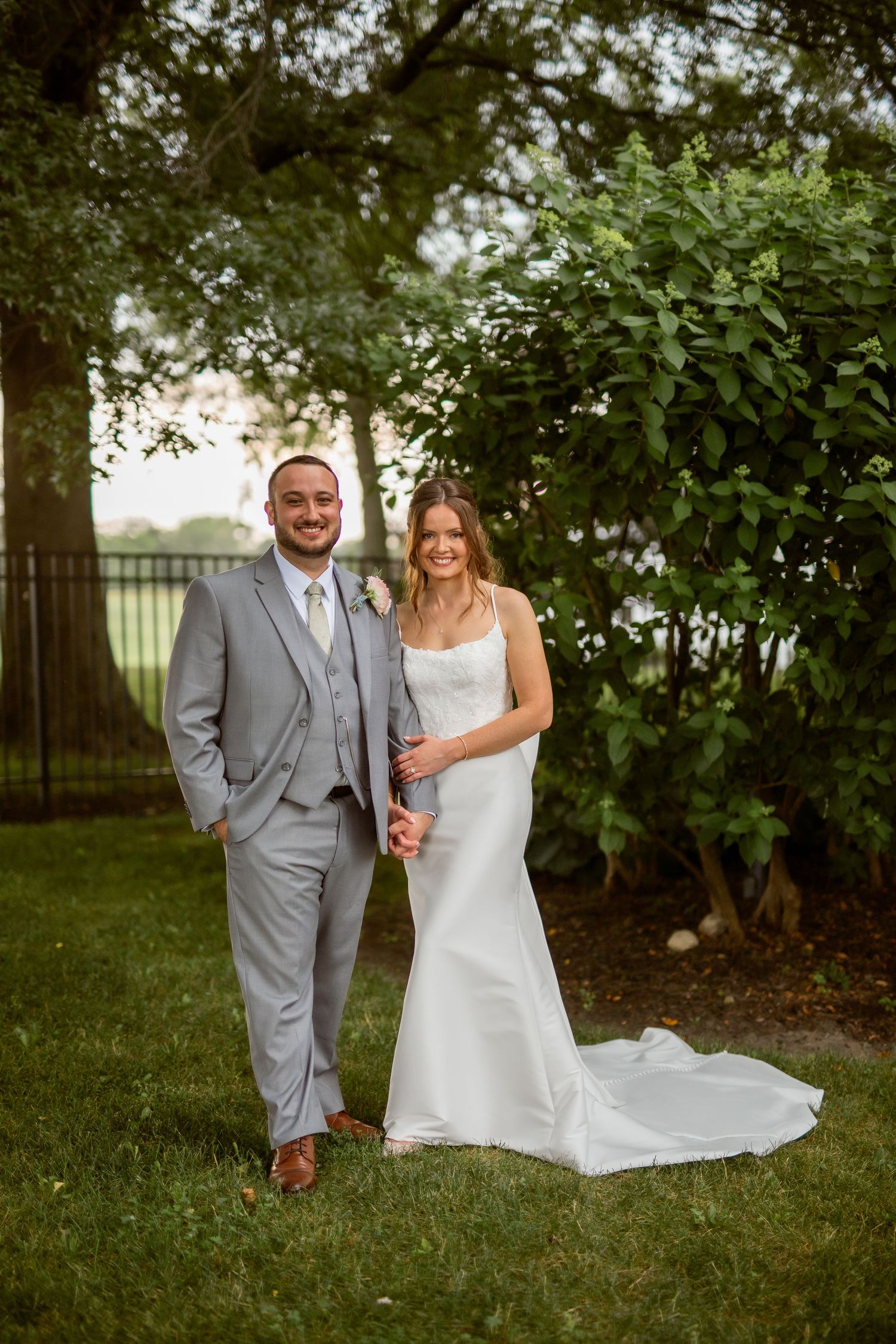 A bride and groom pose together outdoors on grass, she in a white gown, he in a gray suit, with lush trees behind.