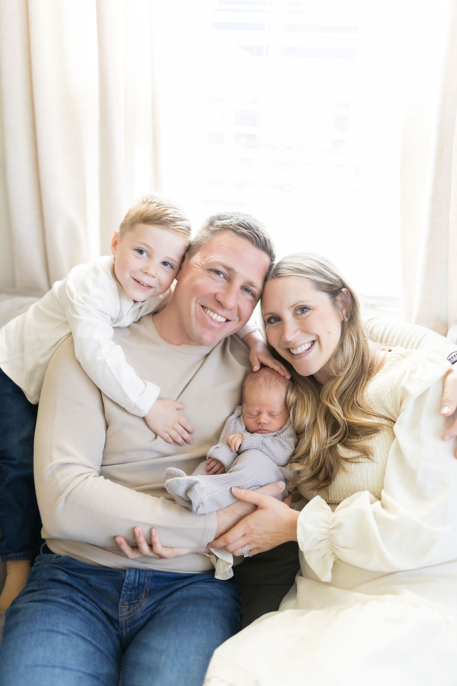 Happy family of four posing together, parents holding newborn baby with young boy smiling behind dad.