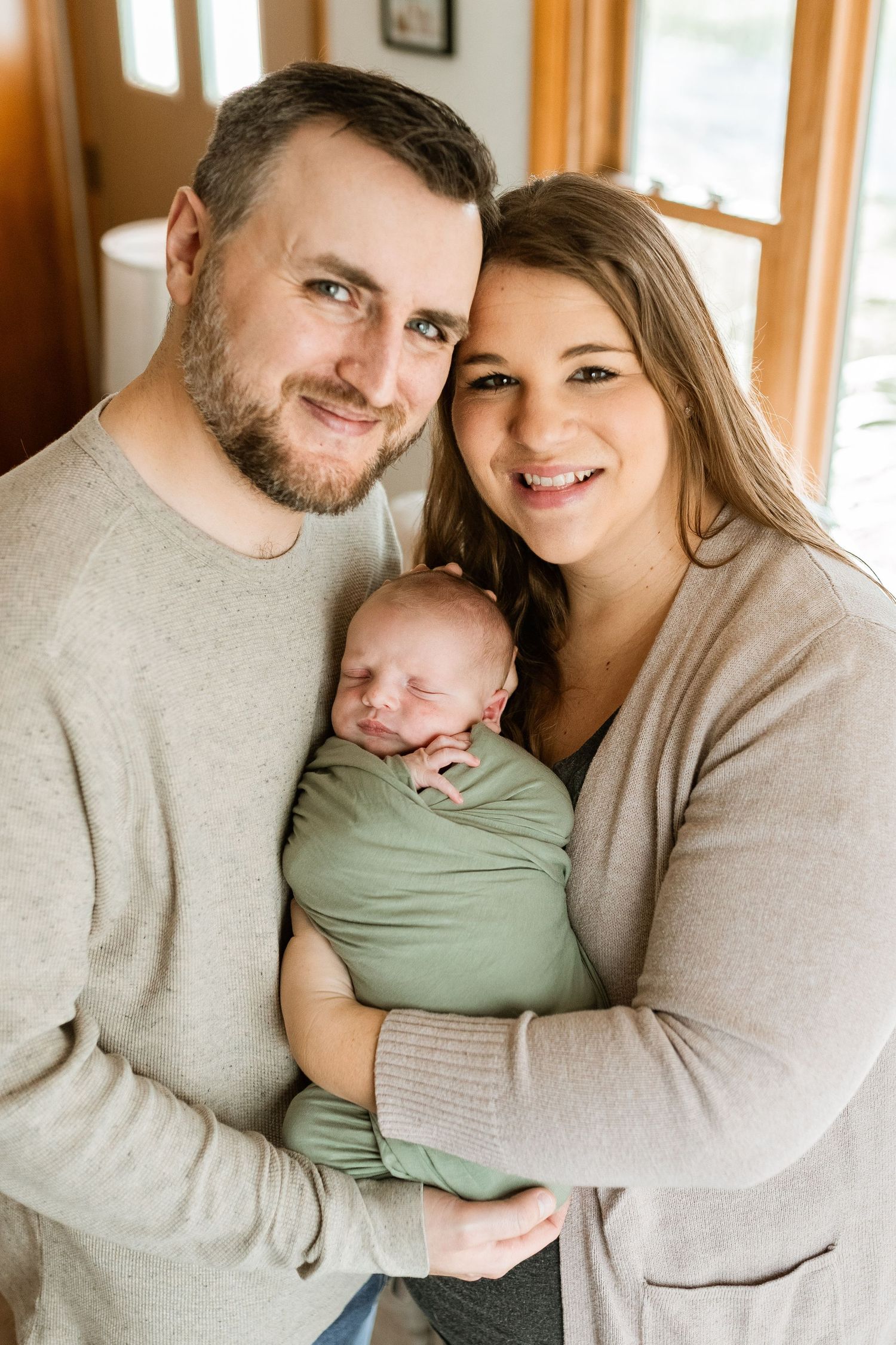 A cozy family portrait shows parents holding their newborn baby in a sunlit room with warm wood accents.