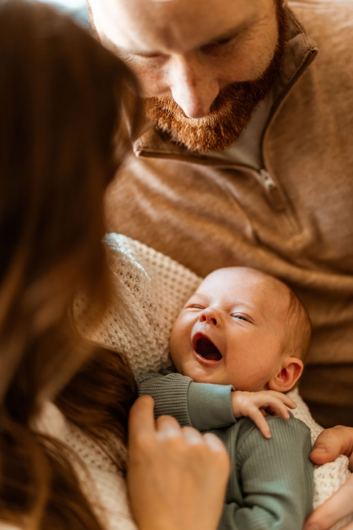 A laughing baby in a mint green outfit lies cradled while being adored by their parents in warm lighting.