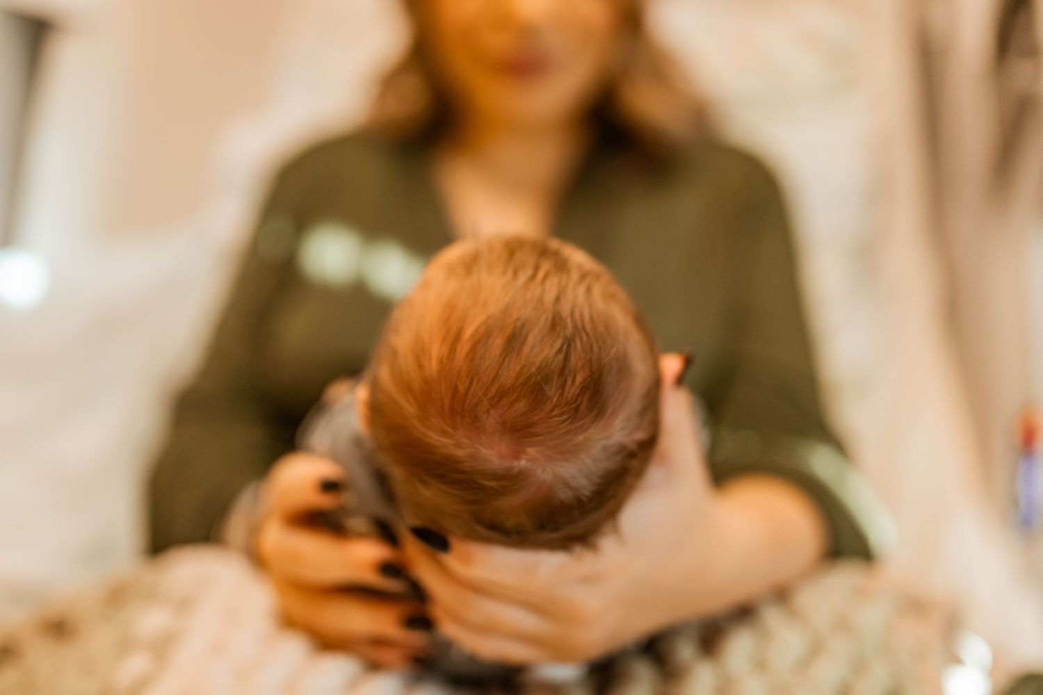 A person holds a small infant with red hair against a soft blurred background in a gentle indoor setting.