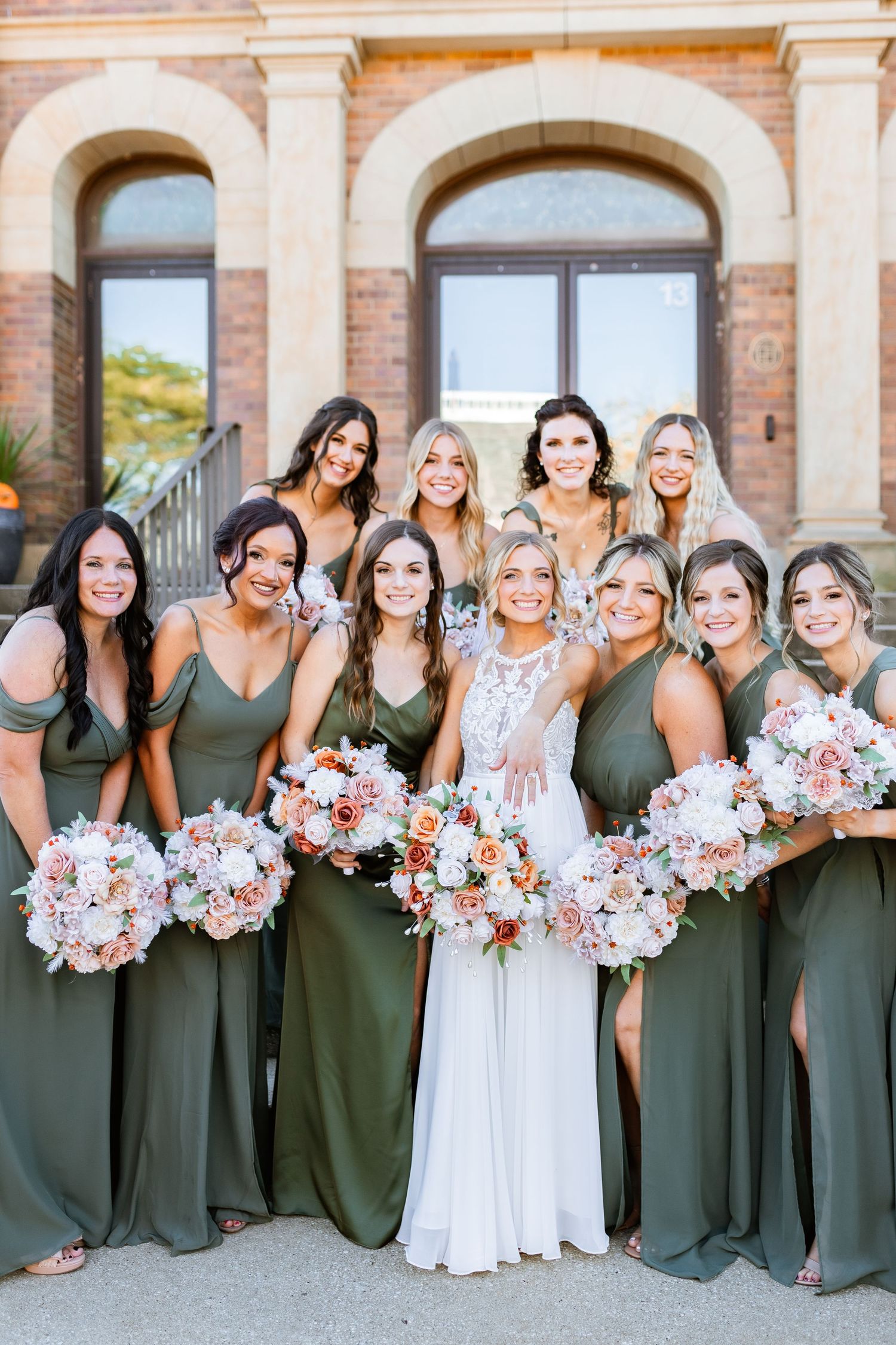Wedding party in olive green bridesmaid dresses holding pink and white bouquets pose with bride outside stone building.
