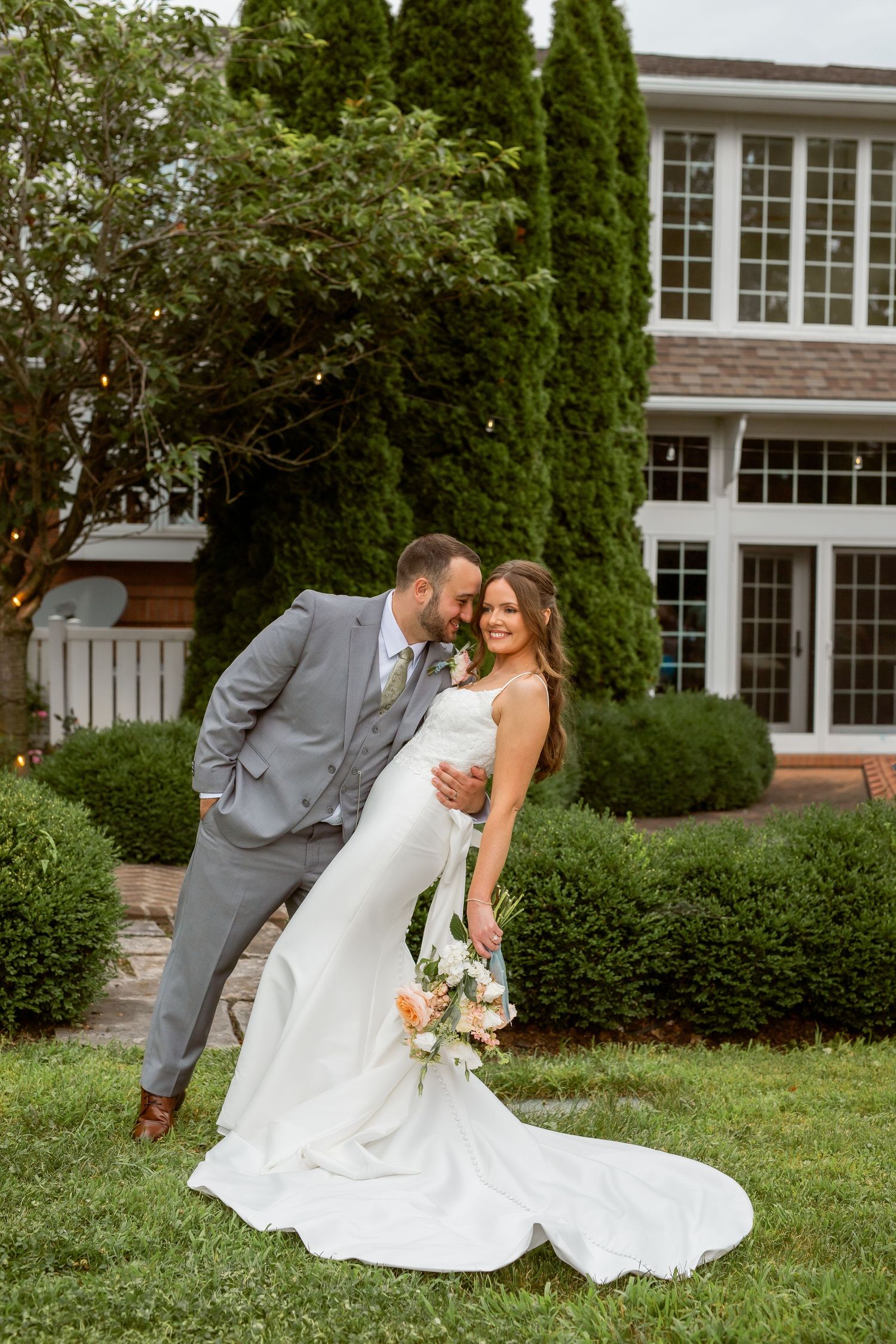 A bride and groom pose together in a lush garden outside an elegant venue, she holds a floral bouquet in her white gown.