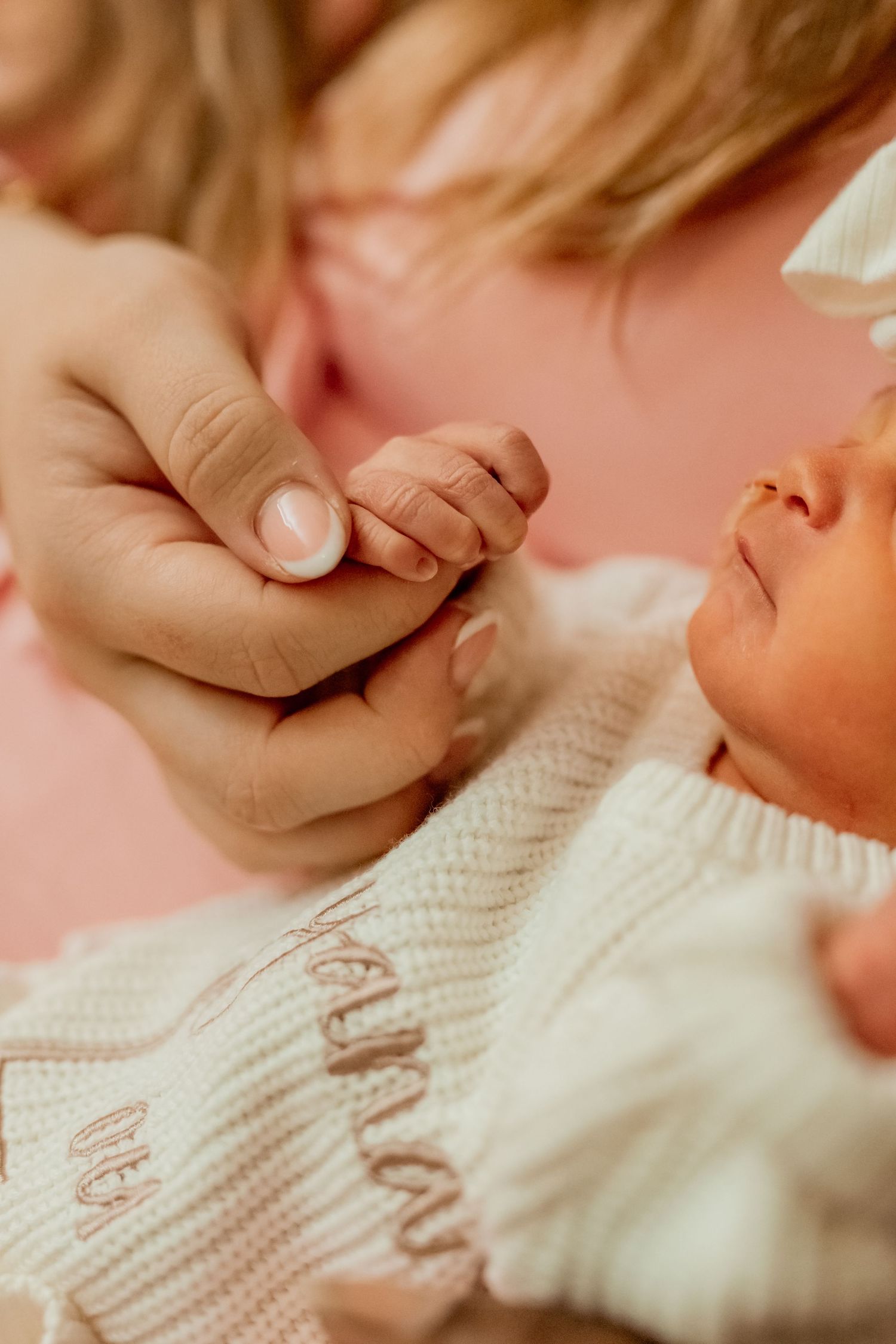 A tiny baby's hand grasps an adult's finger while wearing a white knitted sweater against a soft pink background.