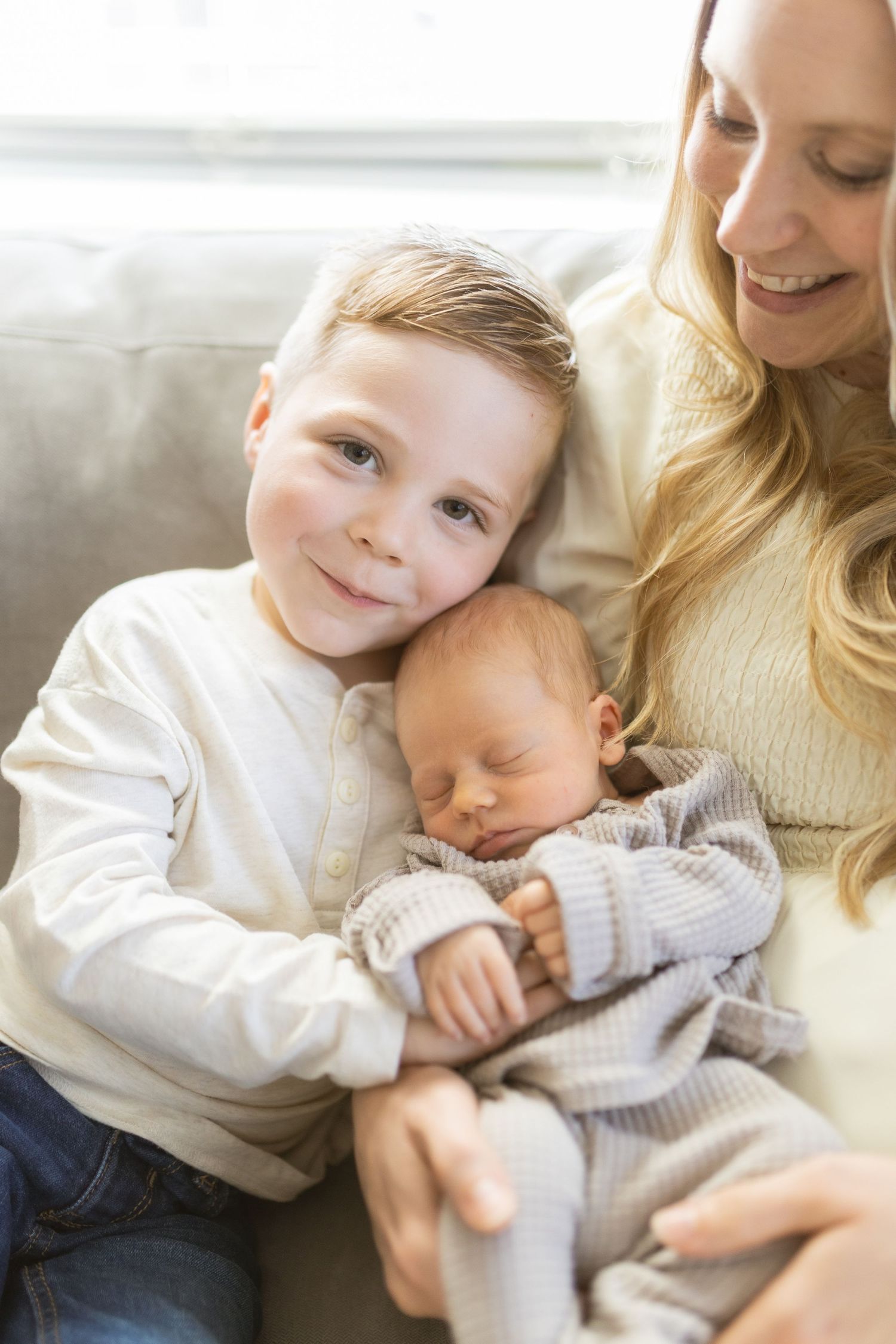 Young boy smiling while holding sleeping newborn baby sibling, sitting with blonde mother on couch.