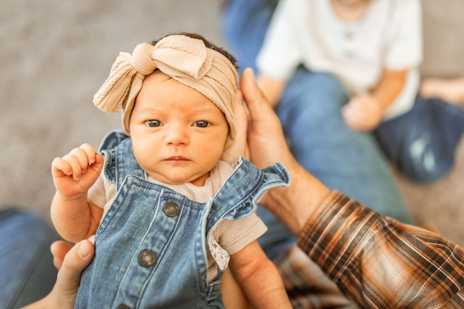 A baby wearing denim overalls and a beige beanie makes playful hand gestures while sitting indoors.