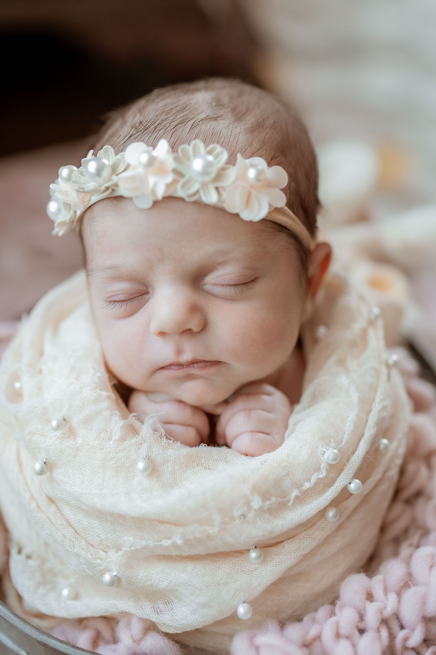 Sweet newborn baby wrapped in cream fabric with pearl-adorned floral crown resting peacefully.