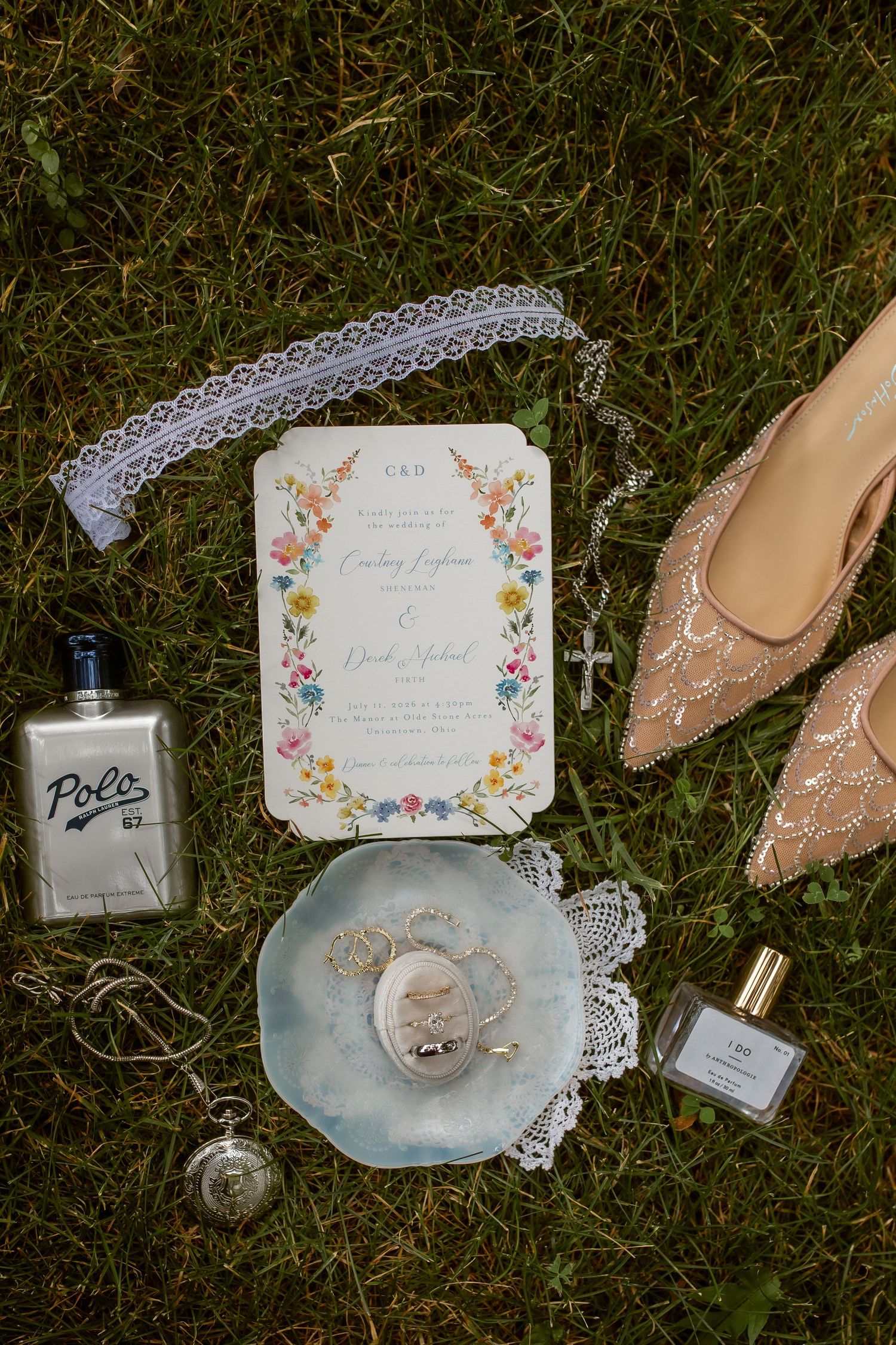 Wedding flat lay on grass featuring floral invitation, nude heels, locket, Polo cologne, perfume, and lace garter.