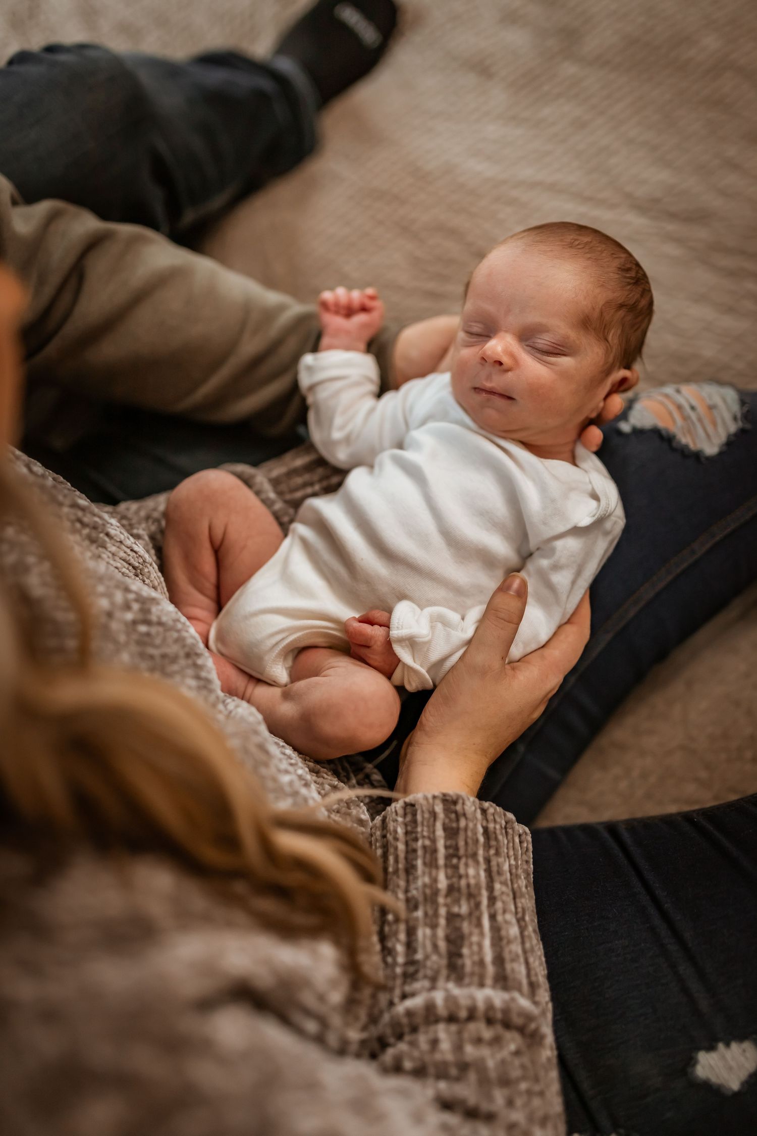 Tiny newborn baby in white onesie resting peacefully on soft blanket while caregiver gently touches.
