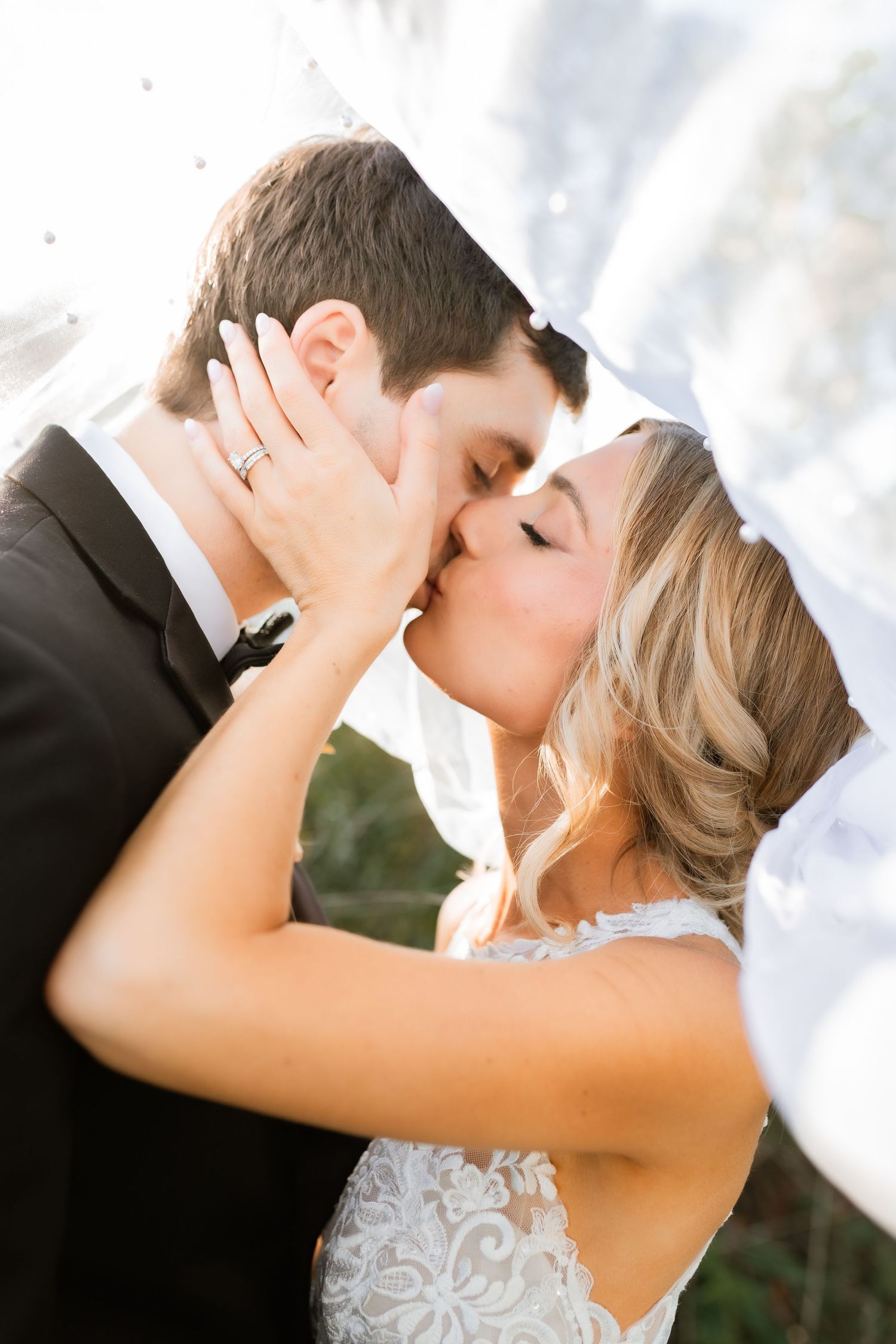 Romantic outdoor wedding photo of newlyweds sharing a tender kiss under a flowing white veil.