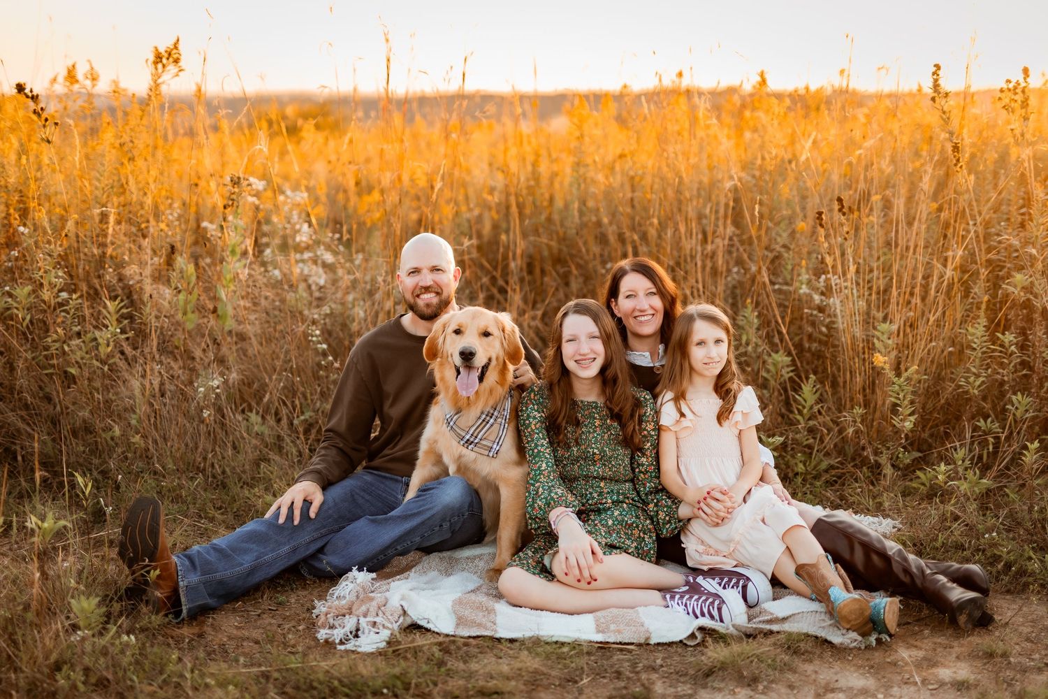 Family of four with golden retriever sitting on blanket in golden field at sunset.
