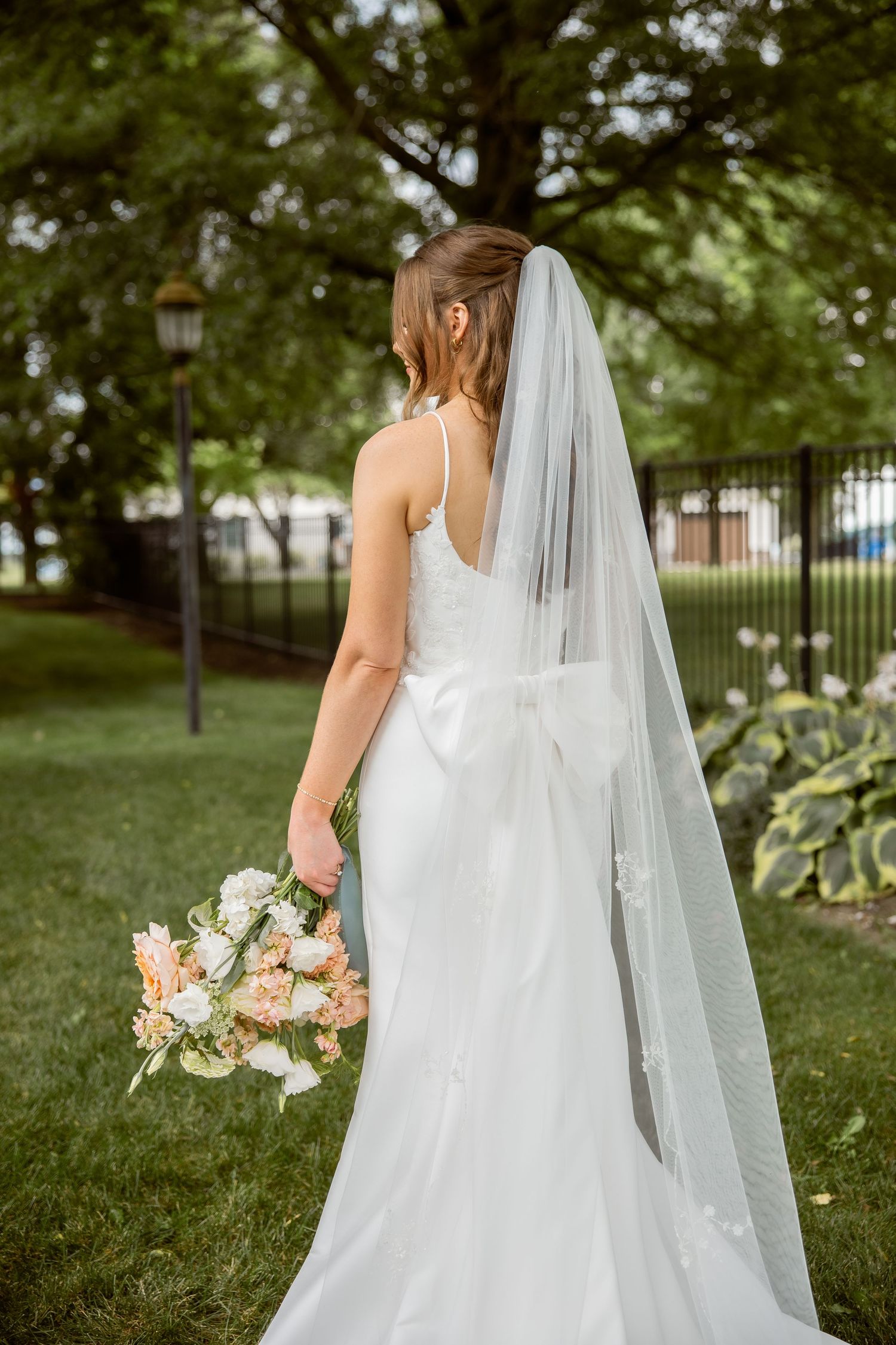 Bride in white gown with long veil holds floral bouquet, standing in lush garden with black iron fence.