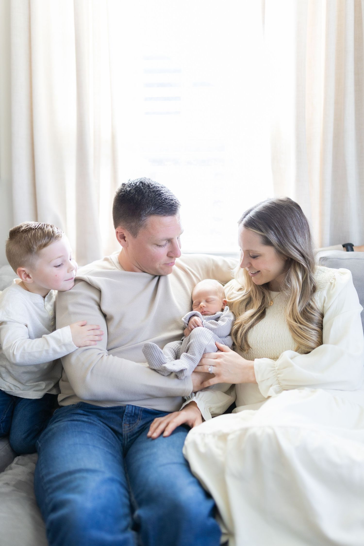 A young family of four sits together on a couch, parents lovingly gazing at their newborn baby with toddler son beside them.
