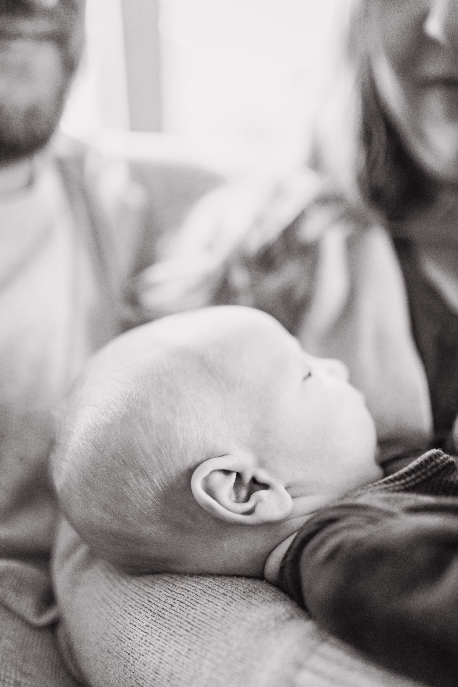 Black and white close-up profile view of an infant's head and ear resting peacefully while adults sit in background.