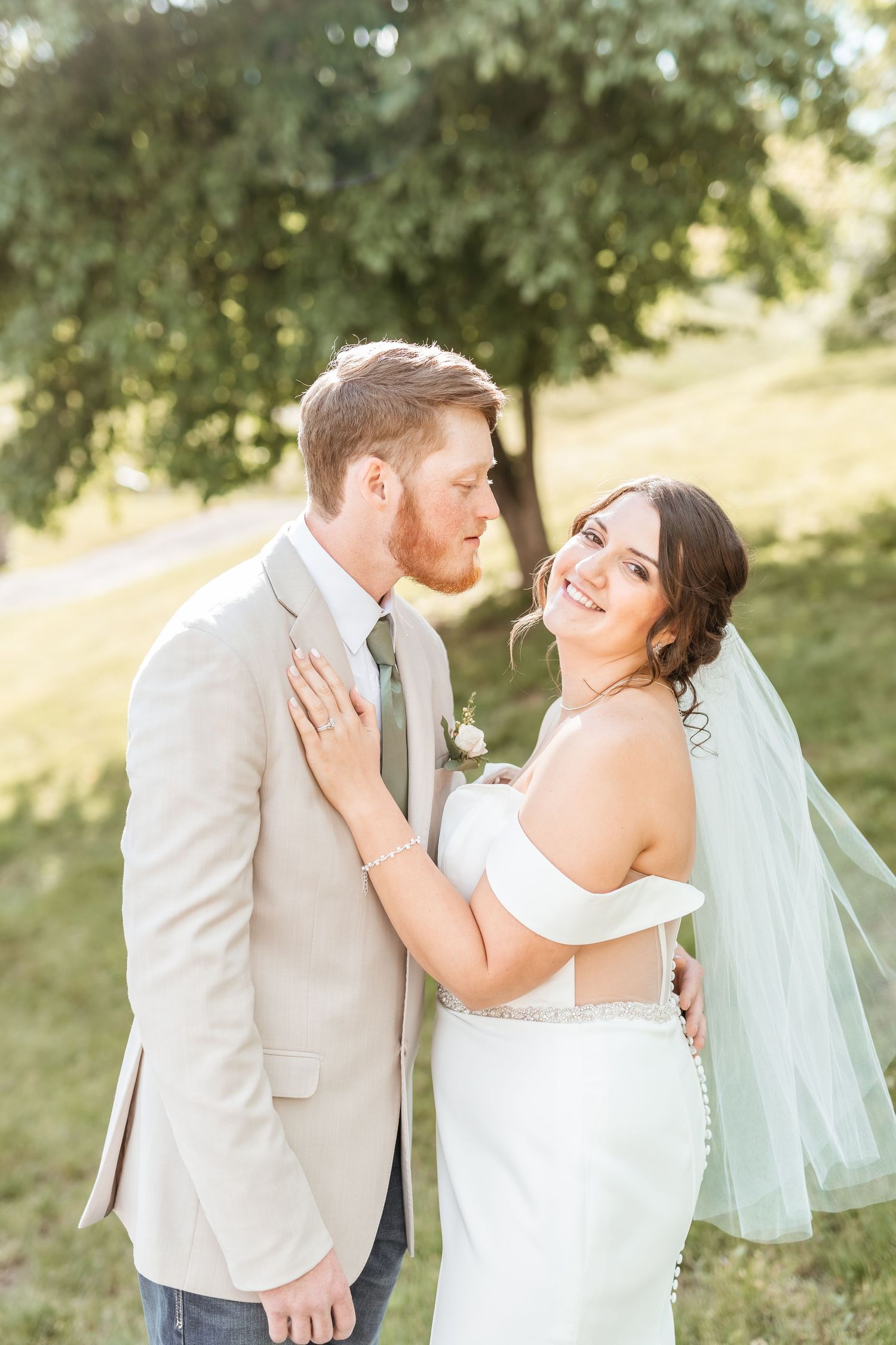Bride in white off-shoulder gown with mint veil smiles as groom in tan suit kisses her cheek outdoors.