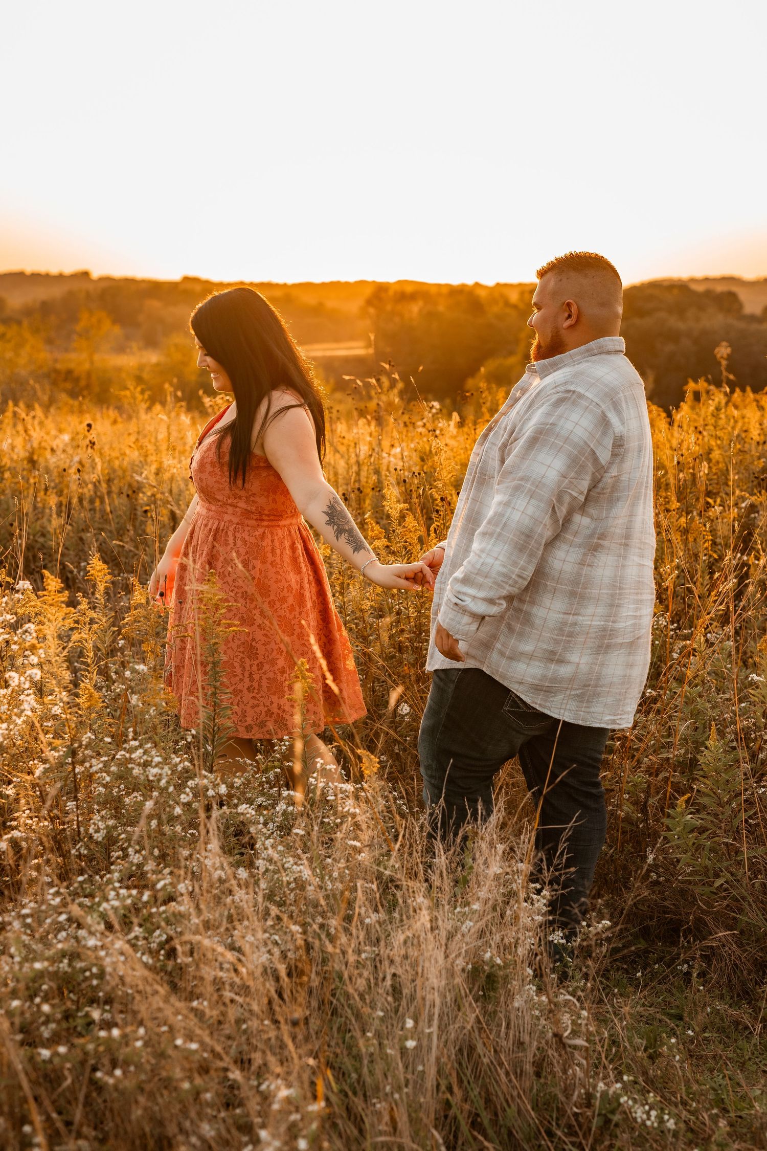 A couple holds hands while walking through a sunlit golden field during sunset.