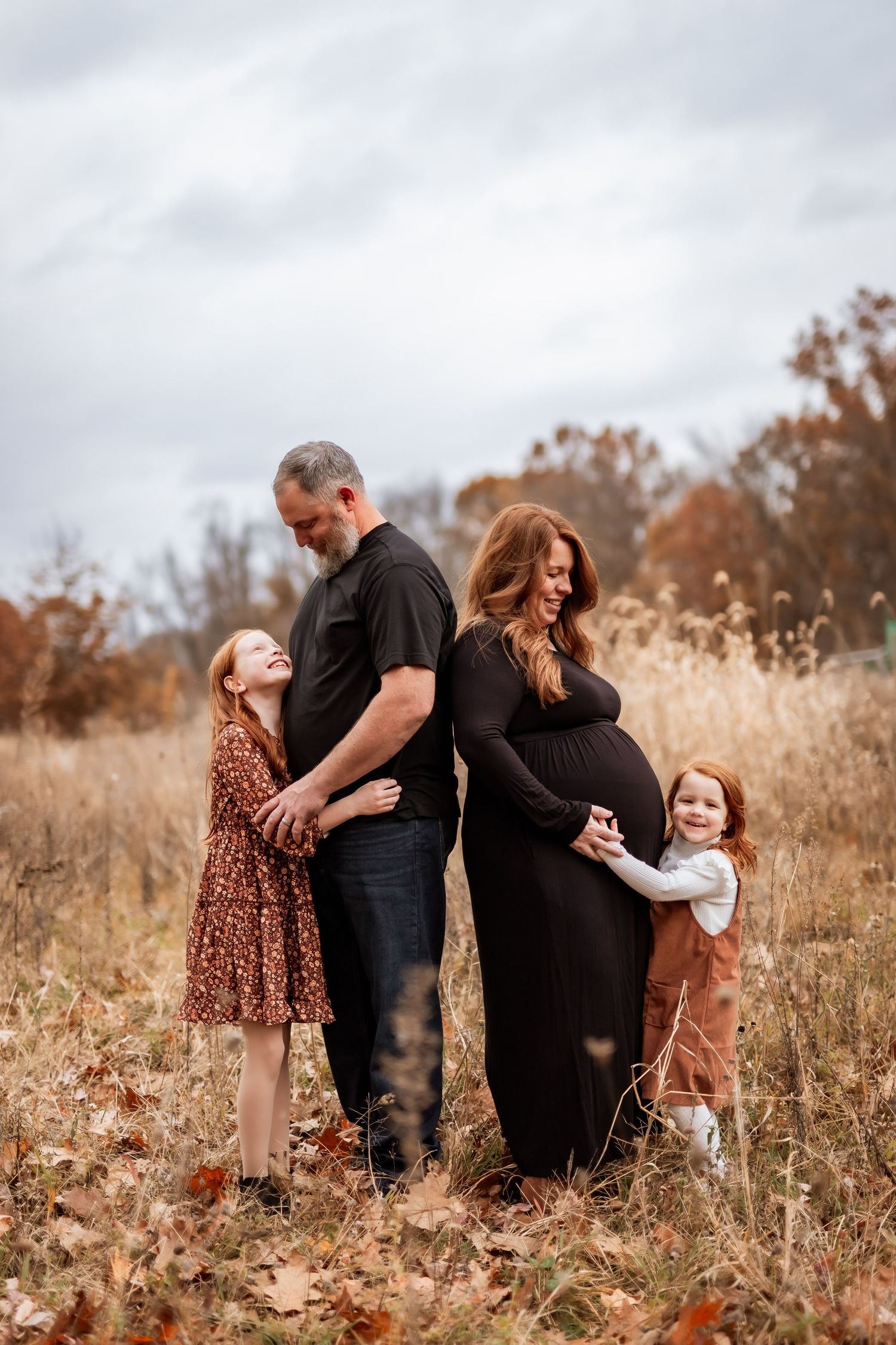 Family of four stands together in an autumn field with tall grass and warm fall colors.