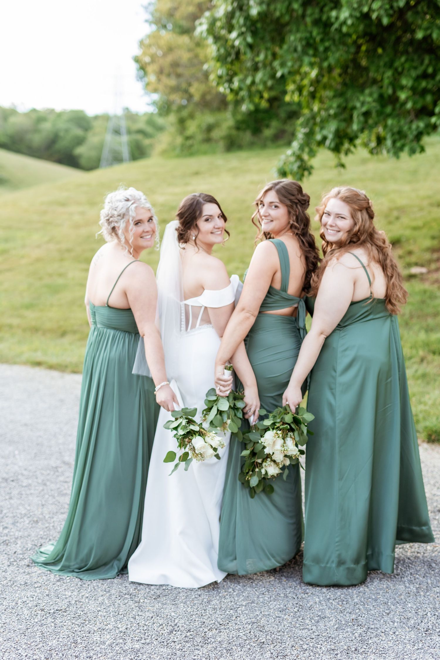 Bridesmaids in sage green dresses sit together and laugh on a gravel path at a rustic outdoor wedding venue.