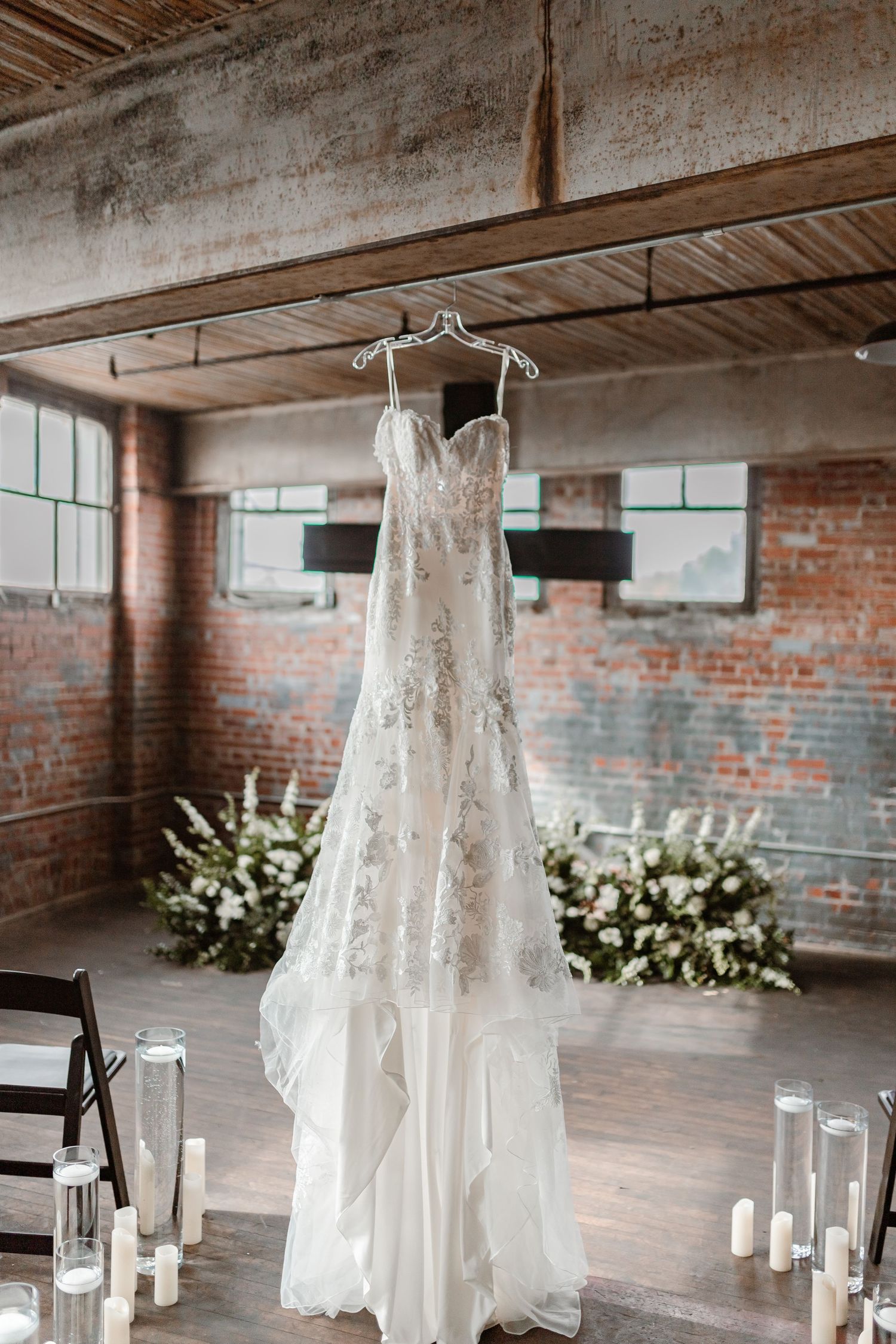 Beautiful lace wedding dress hangs from exposed beams in rustic industrial venue with candles and white floral arrangements.