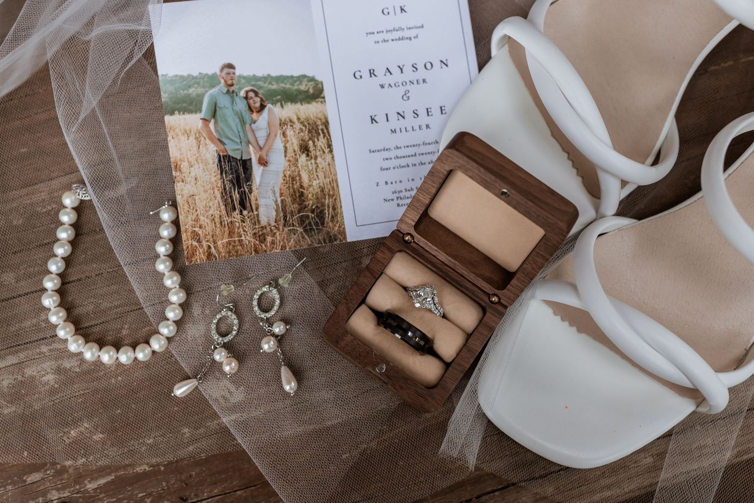 Wedding details flatlay with pearl necklace, earrings, rings in wooden box, invitation card and white shoes on wooden surface.