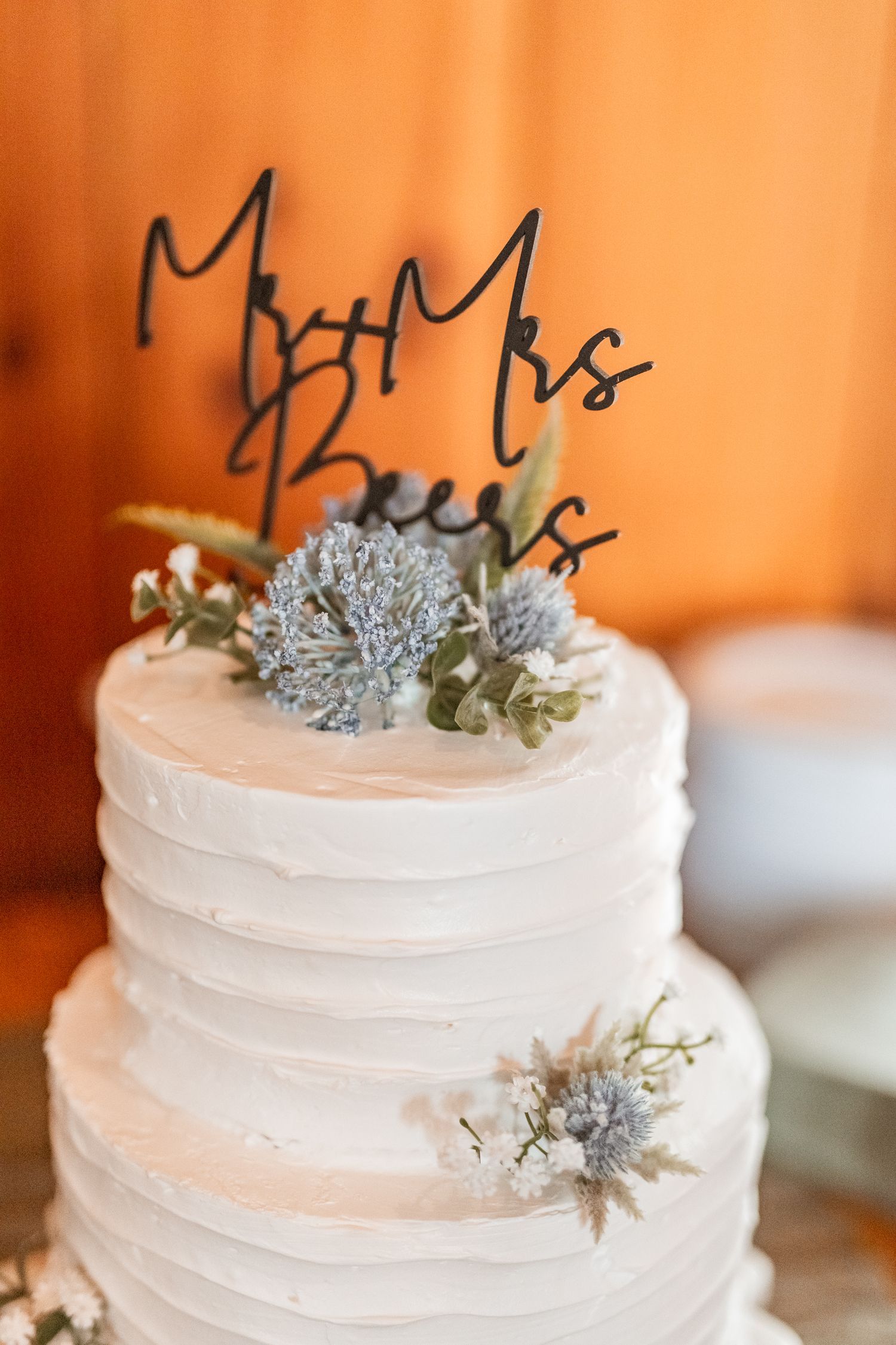 Two-tiered white wedding cake with textured buttercream frosting, delicate blue thistle flowers, and a black cake topper.