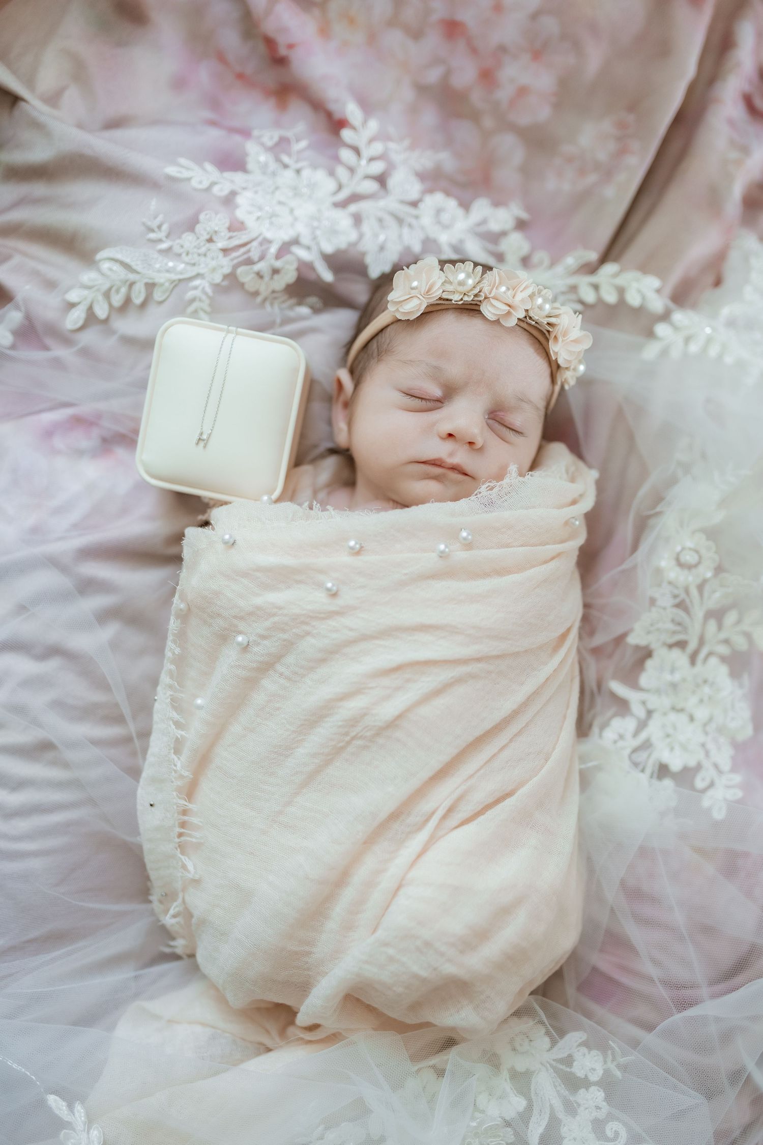 A sleeping newborn baby wrapped in soft cream-colored swaddle blanket lies on delicate white lace fabric.