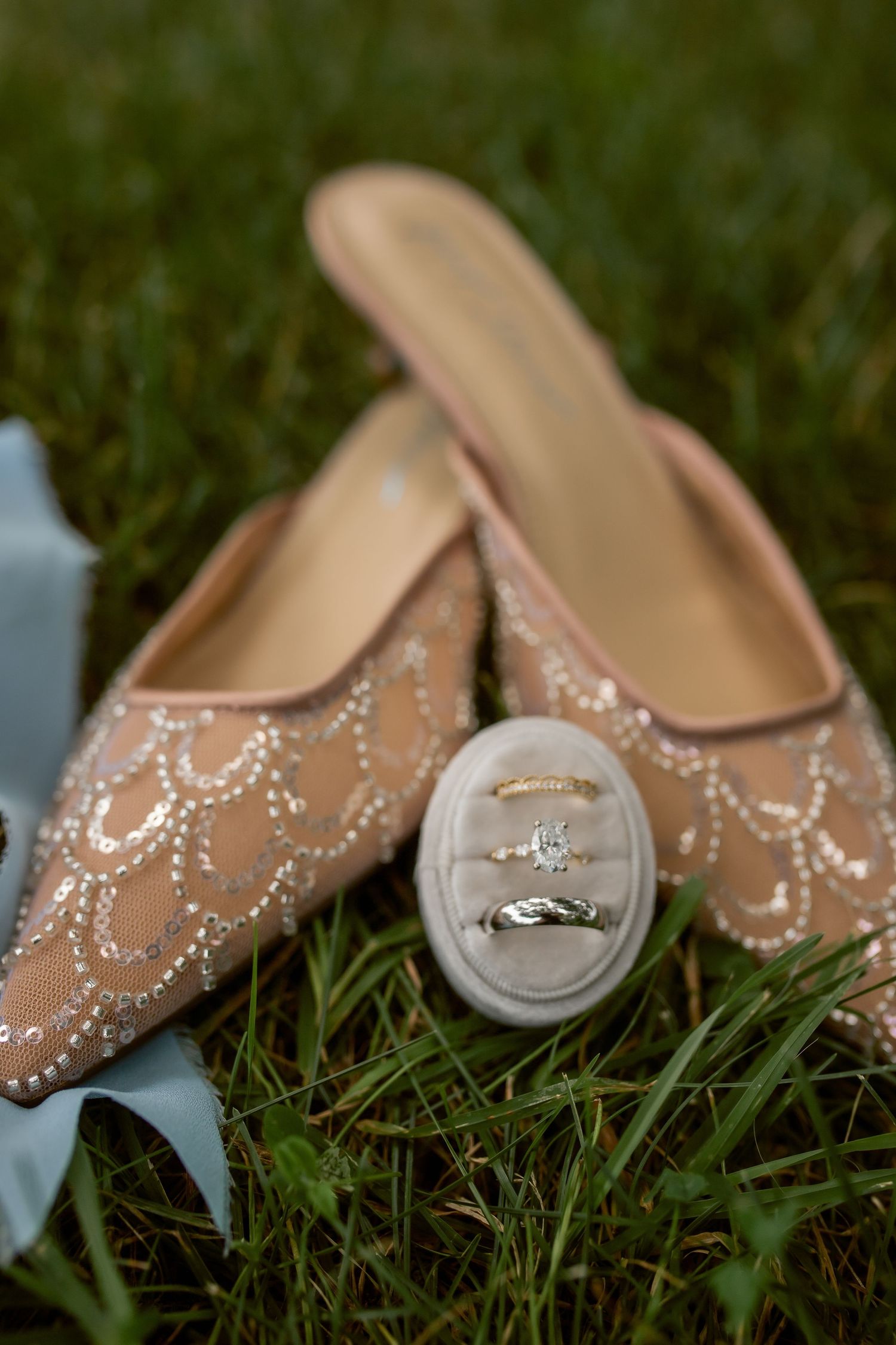 Elegant beaded blush wedding shoes on grass with diamond engagement ring and gold wedding band on a velvet ring holder.