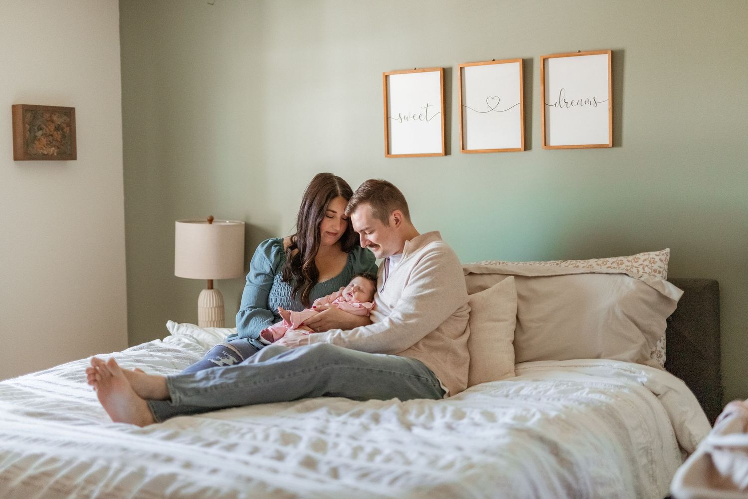 A couple sits on a bed admiring their newborn baby in a softly decorated bedroom.