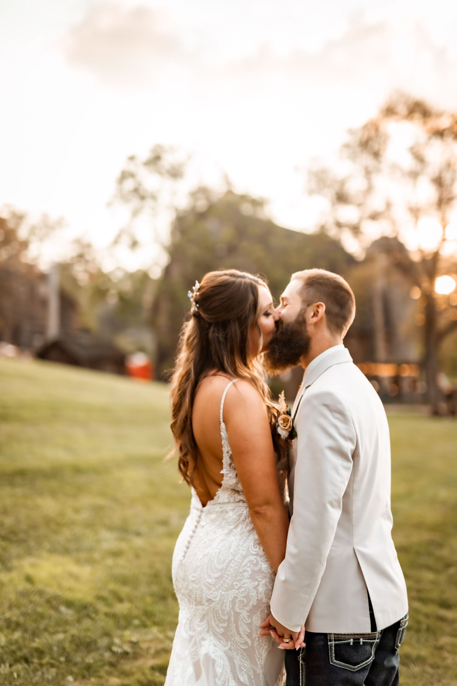 Bride and groom sharing a kiss at golden hour, she in a lace backless gown, he in a white suit.